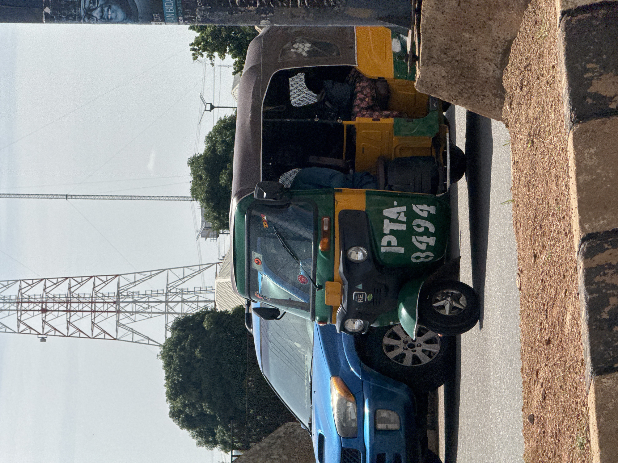 In the city of Bar Jirgi Summa, Nigeria, a green and yellow auto-rickshaw with license plate PTA-8494 is parked on a paved road next to a blue SUV. The auto-rickshaw has a brown roof and a driver wearing a blue shirt, and passengers are visible inside. In the background, a large radio tower stands against a bright sky, with trees and some buildings also present. A portion of a political poster featuring a man's face is visible on a pole to the right. The foreground shows a dirt embankment with a concrete curb.