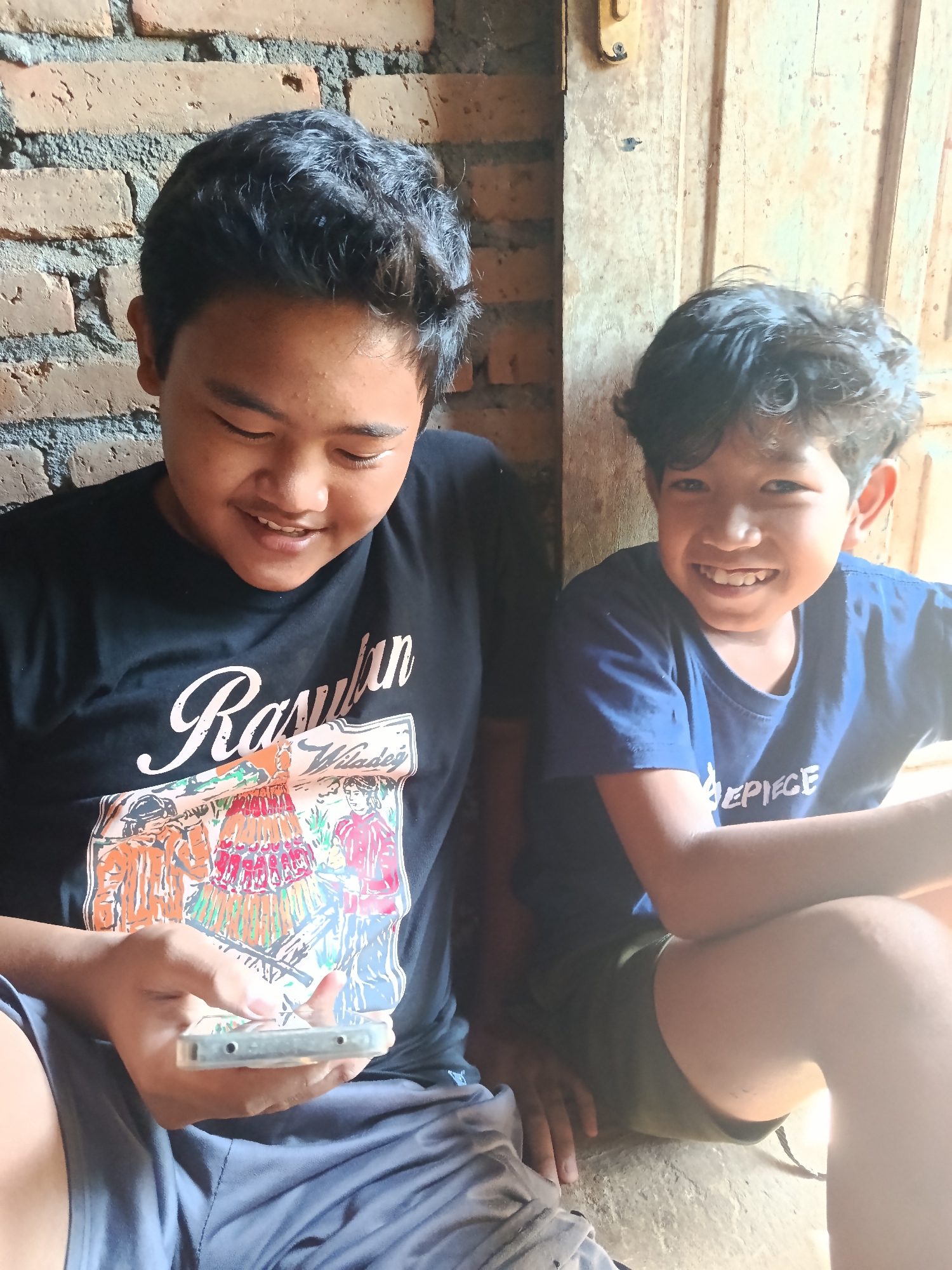 Two young boys are sitting in front of a brick wall and a wooden door in Karangmojo, Indonesia. The boy on the left, wearing a black t-shirt with a graphic print and grey shorts, is looking down at a smartphone he is holding. The boy on the right, wearing a blue t-shirt and dark green shorts, is smiling at the camera. The environment appears to be outdoors or in an open structure, with natural light illuminating the scene.