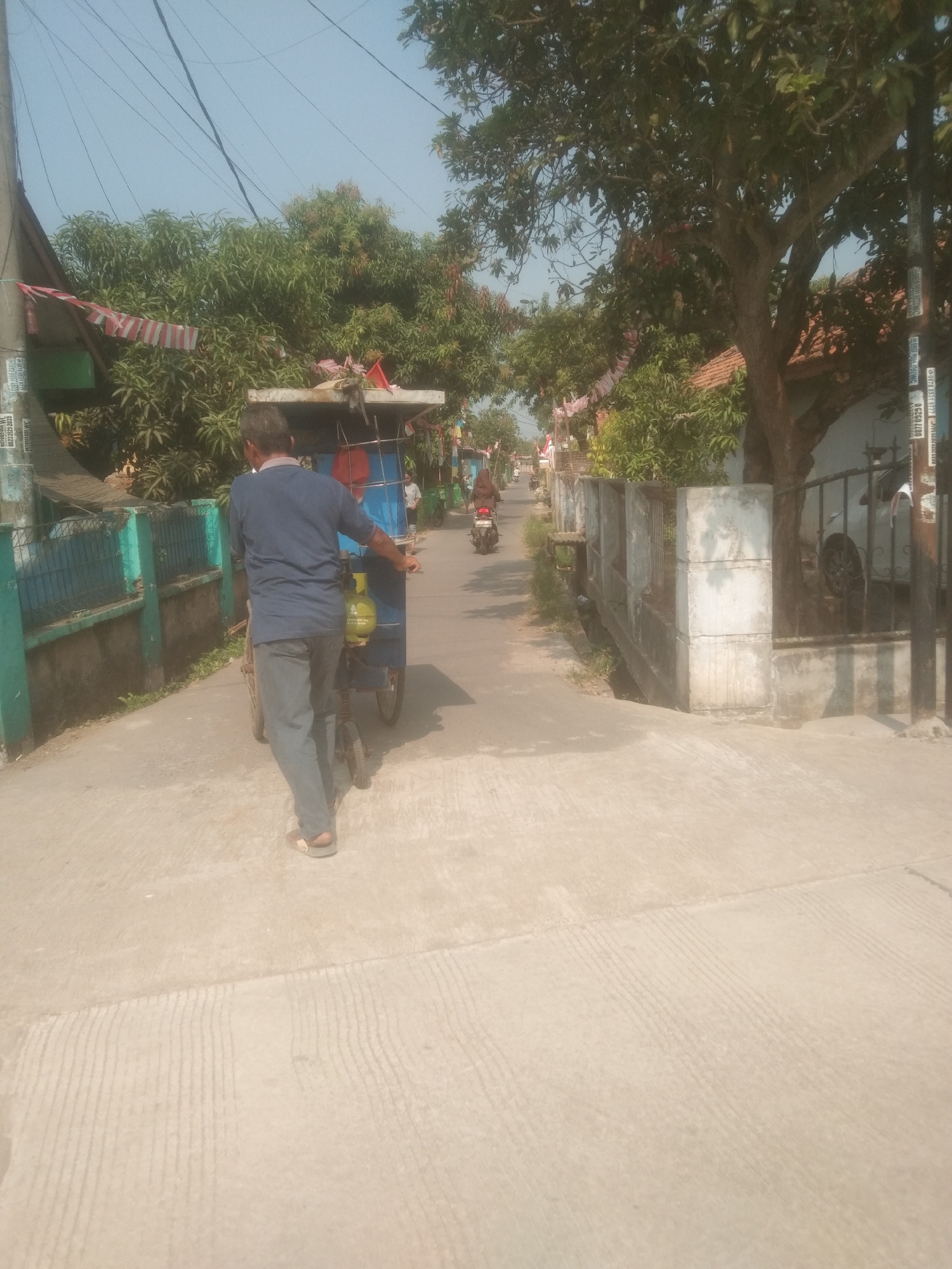 The image depicts a street scene in Subang, Indonesia, under a clear sky. In the foreground, a man in a blue shirt and grey pants is pushing a three-wheeled cart. The cart has a blue body and a yellow gas cylinder attached to it. The man is facing away from the camera, walking down a paved street.
The street is lined with houses and trees. On the left side of the street, there is a turquoise fence and some vegetation. On the right side, there is a concrete wall and a tree with a thick trunk. In the background, other people can be seen on motorcycles, and more houses and trees are visible. Red and white striped flags are hanging from some of the trees, possibly indicating a local celebration or event. The overall impression is a typical residential street in a tropical climate.