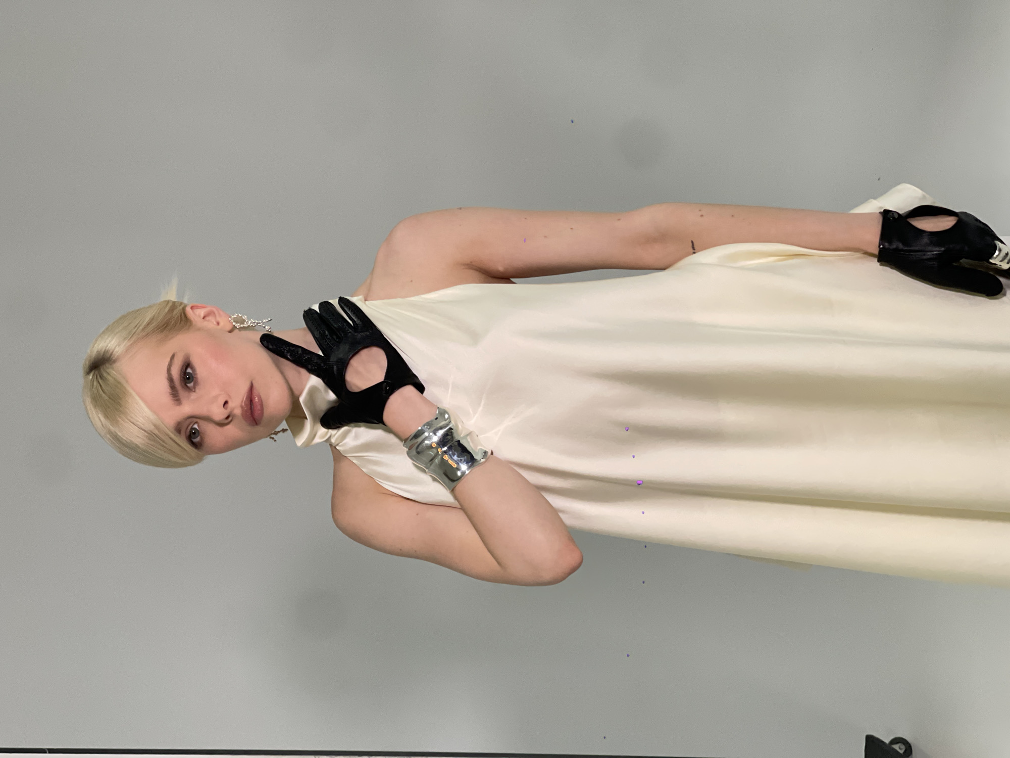 A blonde woman with short hair stands against a plain gray backdrop. She is wearing a sleeveless, off-white dress with a high neckline. Her left hand, adorned with a large silver cuff bracelet, rests on her chest, gloved in black leather. Her right hand, also in a black leather glove, is raised to her face, with her index finger pointing upwards. She is wearing statement earrings. The image appears to be taken indoors in a studio setting, likely in Kyiv, Ukraine.