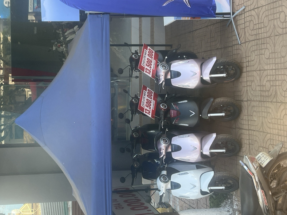 The scene depicts an outdoor commercial display of electric scooters situated on a tiled sidewalk in Trung Hồ, Vietnam. A blue pyramidal canopy tent covers eight VinFast EVO Lite Neo electric scooters arranged in two rows. The front row, from left to right, features one white, two light pink/lavender, and one dark gray/black scooter. Two red signs displaying "EVO LITE NEO 13.500.000" are attached to the handlebars of the two light pink scooters. A partially visible white sign on the far left reads "GIÁ TỪ 1.000.000". In the lower left foreground, a silver and black Yamaha Sirius motorcycle with a partially legible license plate "17-K8 102.XX" is parked on the patterned tiled ground. The background reveals a commercial storefront with large glass windows reflecting ambient light and elements from across the street. A blue vertical banner on a stand is partially visible on the right. No individuals are present in the image. The environment is under bright daylight conditions.