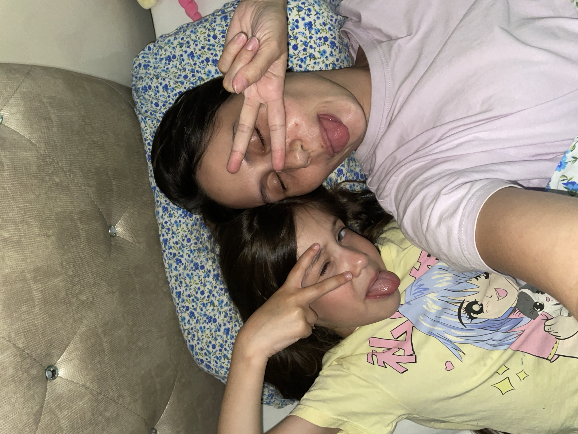 Two individuals, an adult and a child, are lying on a bed. The adult is wearing a light pink t-shirt, while the child is wearing a yellow t-shirt with an anime graphic. Both are making a peace sign with their fingers over their eyes and sticking out their tongues. They are lying on a bed with a blue floral patterned pillow and a grey headboard with diamond-shaped decorations. The photograph appears to be taken indoors. The location is not discernable from the image, so stating it is in Izhevsk, Russia is unsubstantiated.