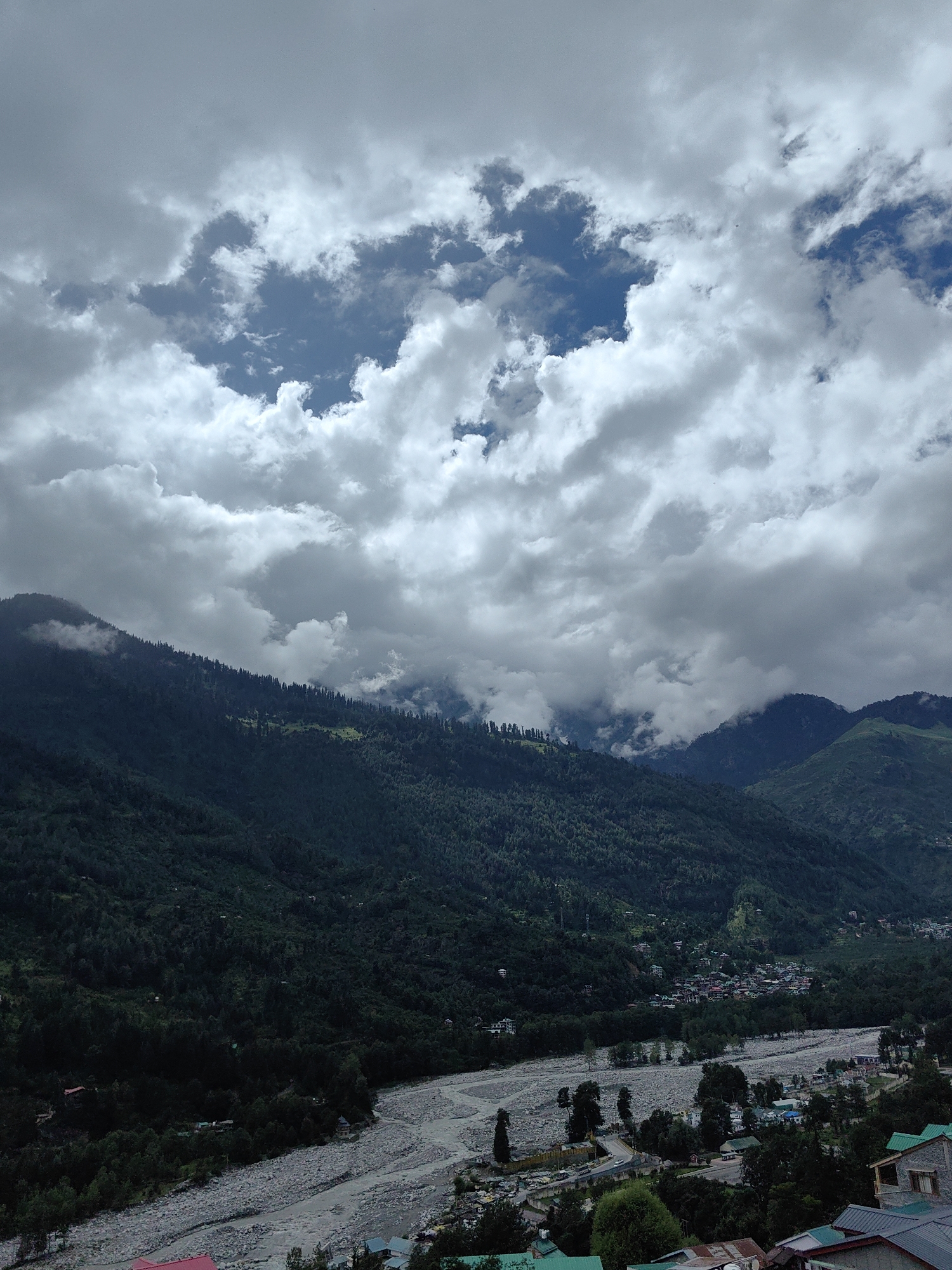 The image depicts a panoramic view of a mountainous landscape with a river flowing through a valley. The sky is filled with dramatic, heavy clouds, with patches of blue visible through the gaps. The mountainsides are densely covered with dark green trees, and in the distance, a small town or village is nestled within the valley. The riverbed appears to be wide and stony, with the water flowing through it. The overall impression is one of a vast and natural environment, with human settlement present but dwarfed by the scale of the landscape.
The image captures a scene in the city of Vashisht, India, renowned for its hot springs and its location in the Kullu Valley of Himachal Pradesh. The mountainous terrain, dense forests, and the presence of a river are characteristic features of this region. The cloudy sky suggests a possibility of monsoon season or overcast weather, which is common in the Himalayas. The village or town visible in the valley is likely a settlement that has developed along the river, utilizing the natural resources of the area. The architecture of the buildings, though not clearly detailed due to the distance, appears to be typical of rural or semi-urban settlements in the region, possibly with pitched roofs to withstand snow. The image conveys a sense of serenity and natural beauty, highlighting the majestic mountain ranges and the life-sustaining river that flows through them.