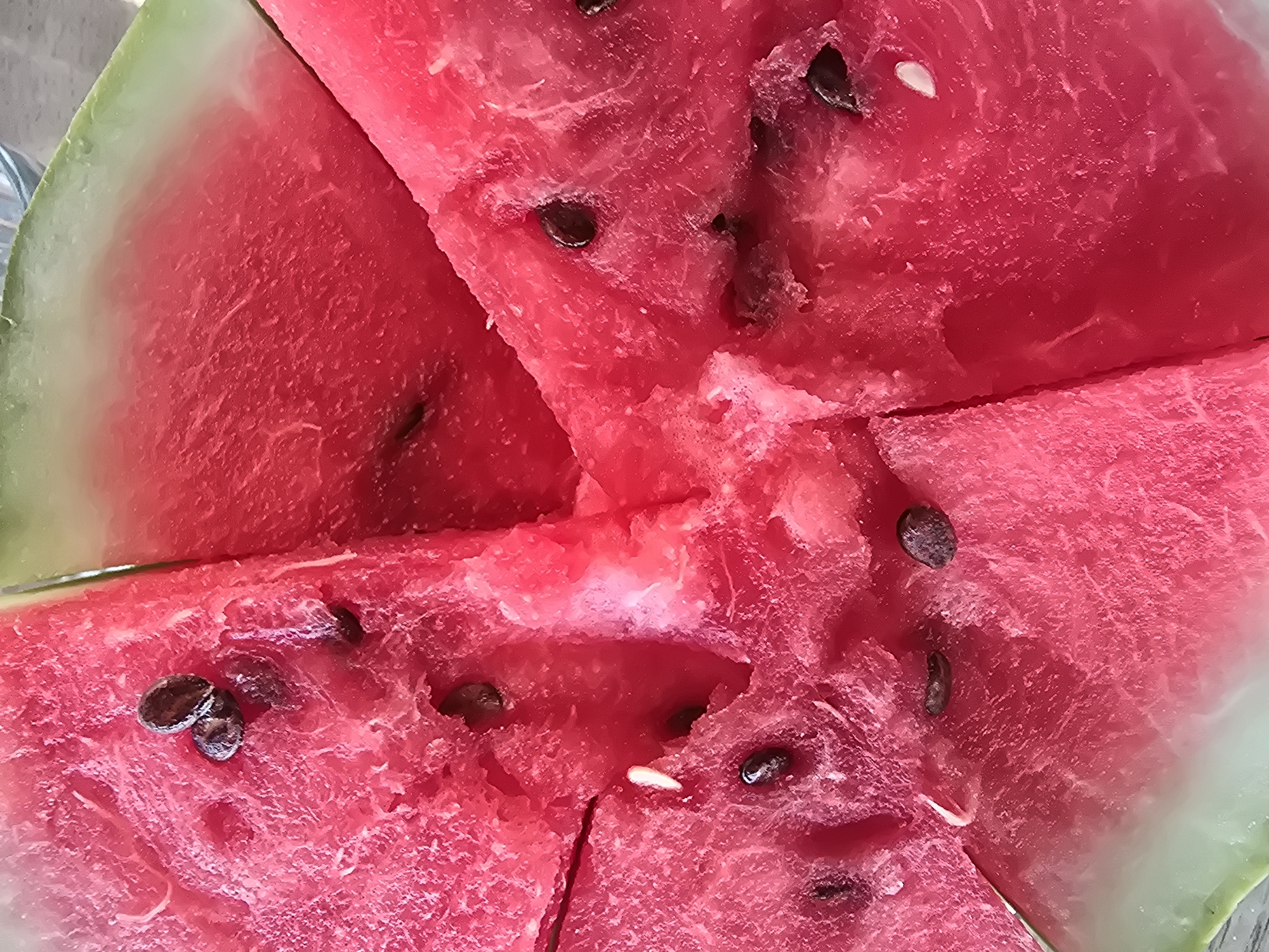 The image displays a close-up, overhead view of several slices of watermelon. The watermelon is a vibrant red with visible seeds, some of which are dark brown and oval-shaped, while others are small and white. The texture of the watermelon flesh appears slightly granular and juicy. One slice shows a gradient from the deep red flesh to a pale green rind, indicating the outer skin of the fruit. The lighting suggests an outdoor setting, possibly natural daylight. There are no people or buildings visible in the frame, and the image does not provide any information about the location of Maksimiha, Russia.
