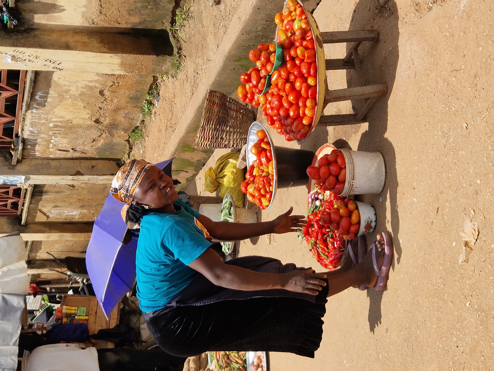 A woman is bent over at a street market, arranging vegetables. She is wearing a bright blue shirt, a long black skirt, and purple flip-flops. Her head is covered with a patterned scarf. In front of her are several large bowls and baskets filled with red tomatoes and chili peppers. A blue umbrella is open behind her. The ground is dusty earth, and a weathered concrete wall is visible in the background. There are other stalls and goods, including sacks and baskets of produce, also present.