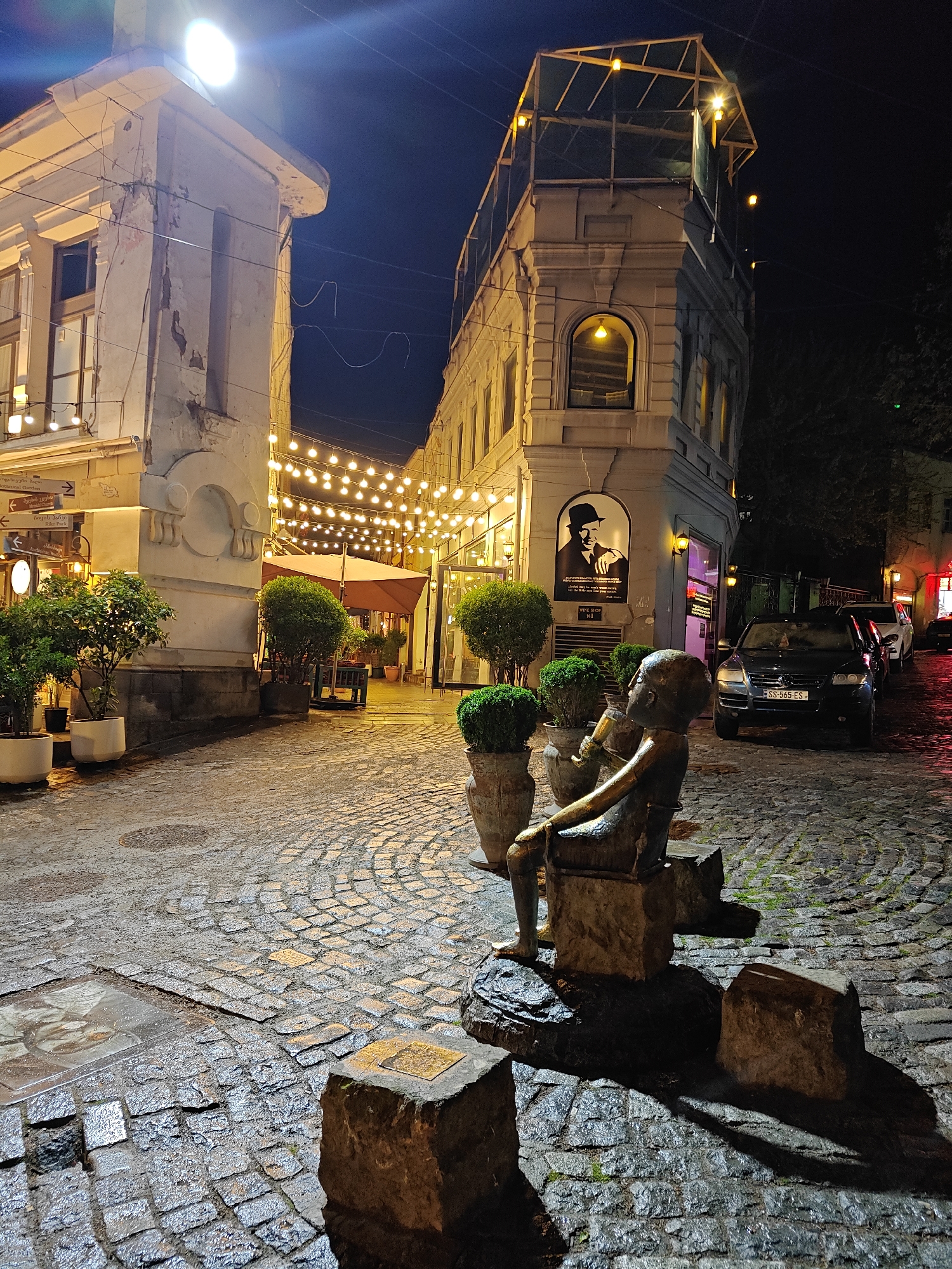 This night-time image presents a charming, wet cobblestone street in Tbilisi, Georgia, bathed in the warm glow of numerous string lights strung overhead between old buildings. The ground is visibly damp and reflective, suggesting recent rainfall.
In the foreground, a weathered bronze statue of a young figure, possibly a boy, sits on a stone block, holding an object to its mouth while looking towards the right. Several potted, well-trimmed evergreen plants are positioned around the statue and along the sides of the street.
To the left, an ornate, light-colored building with peeling paint features brightly lit windows. A sign on its facade displays directional arrows pointing to "Botanical Garden," "Rike Park," and an "Arabic restaurant," accompanied by Georgian text. Further down this side, an outdoor seating area with a large umbrella is visible under the string lights.
The street curves to the right, revealing a distinct corner building. On its exterior, a prominent black-and-white poster depicts a man in a fedora, resembling Frank Sinatra, with the text "WINE SHOP N1" and an partially obscured quote below. An arched window on this building emits a yellow light. The upper section of the building has an open, skeletal framework, possibly a rooftop terrace, also adorned with lights.
Along the right side of the street, several cars are parked on the wet cobblestones, including a black SUV with a Georgian license plate "SS-565-ES." The overall scene evokes a tranquil yet festive atmosphere in a historic urban setting.
