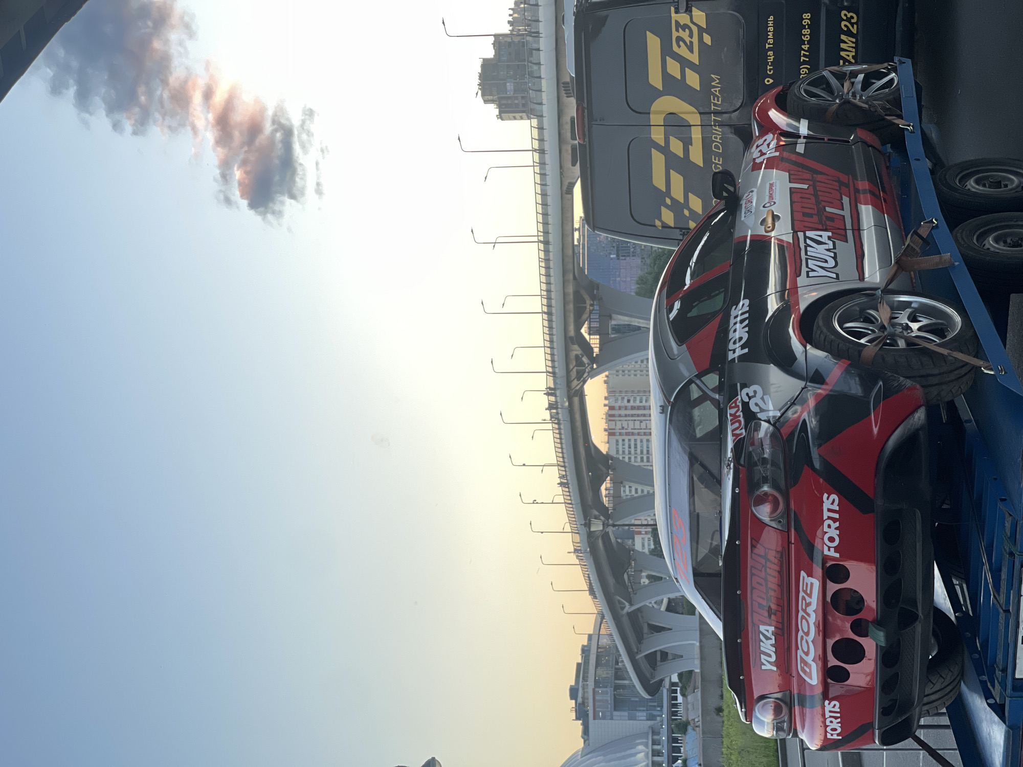 An eye-level, full, exterior shot captures a red and black sports car on a trailer at a highway overpass in an urban setting at dusk. The car, with decals visible, faces right, its rear facing the camera. A black van with yellow lettering is parked behind the car, to the right. The overpass, with streetlights and people walking on it, extends across the frame, forming the background. The sky above is a soft blue with wispy pink clouds. The scene appears to be in a city, though the specific location is not identifiable.