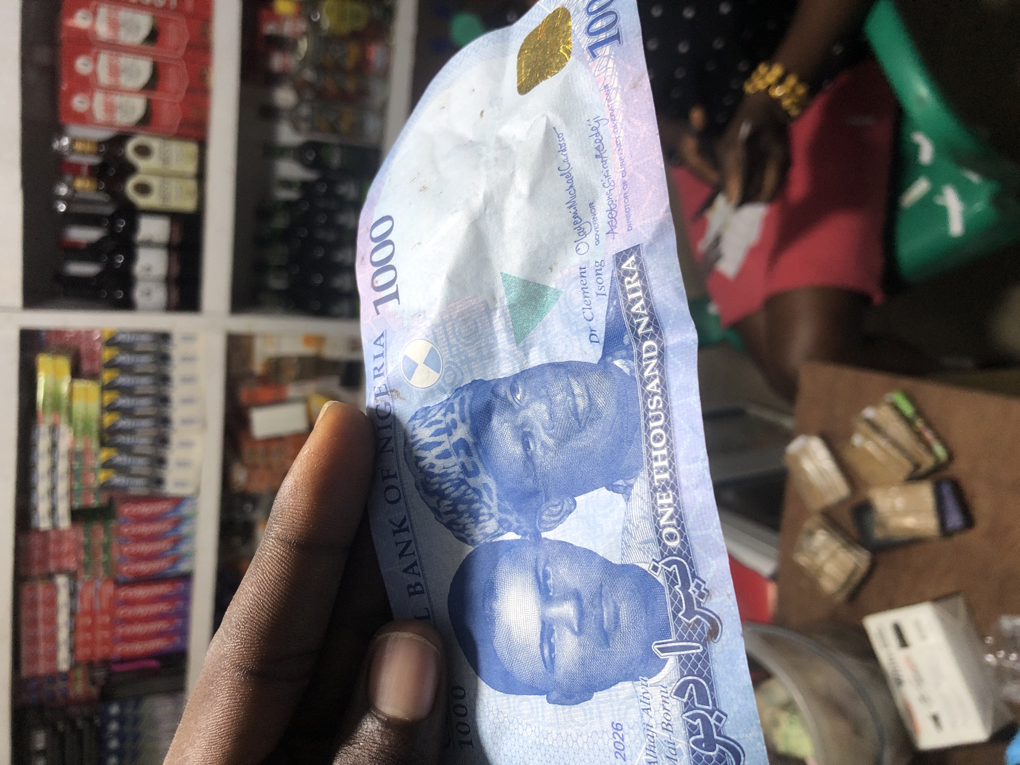 In this close-up image, a Nigerian 1000 Naira banknote is held in a person's hand. The banknote features portraits of two men and text indicating "NIGERIA" and "ONE THOUSAND NAIRA". The background reveals shelves stocked with various items, including what appear to be toothpaste boxes and bottles of liquor, suggesting a retail environment. The lighting is artificial, casting subtle shadows. The focus is on the banknote, with the background slightly blurred.