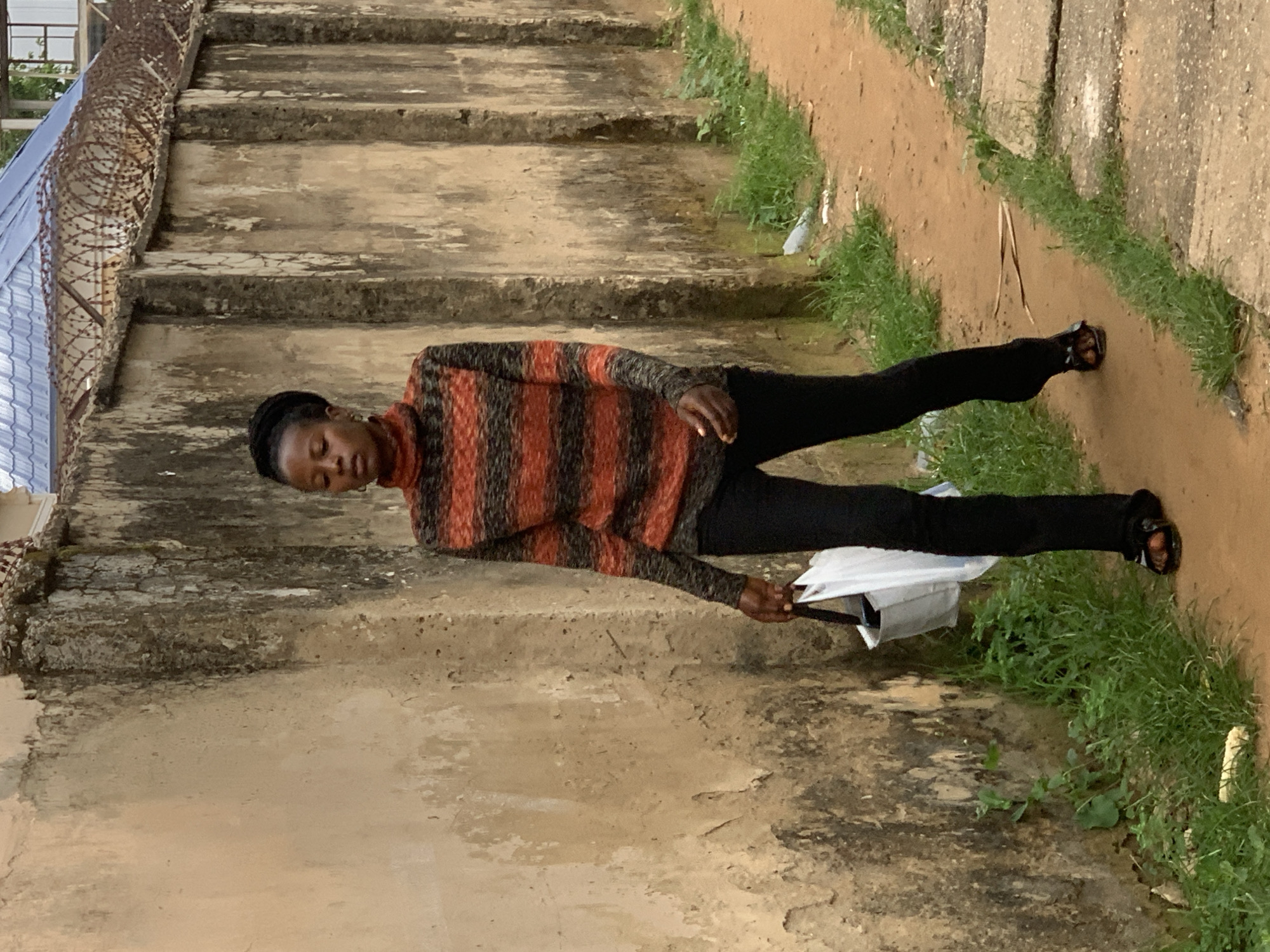 A young Black woman walks along a sandy path next to a weathered, stained concrete wall, on the left side of the frame. She is wearing a striped turtleneck sweater with horizontal bands of orange, red, and black, and black pants. She is carrying a white tote bag in her right hand. Her eyes are closed and her head is slightly bowed. She is wearing black sandals with orange straps. The path is bordered by green grass and some scattered debris. In the background, the wall continues, topped with barbed wire and a portion of a blue roof. This scene is likely located in a residential or less-developed area of Abuja, Nigeria.