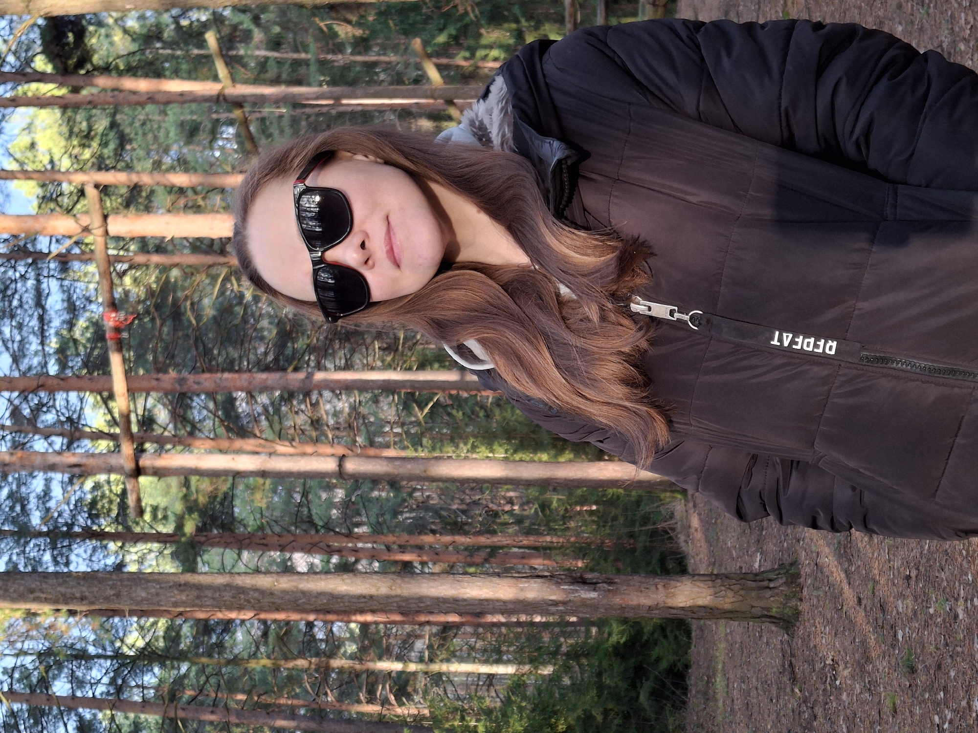 A young woman is depicted posing outdoors in a pine forest, likely in Chaikovsky, Russia. She is shown in a relaxed pose, with her head tilted slightly, and a subtle smile. Her long, wavy brown hair falls over her shoulders. She wears black sunglasses with visible red accents on the frames.
The woman is dressed in a dark, padded winter jacket, which appears to be black or very dark brown. A zipper runs down the front of the jacket, and a black strap attached to the zipper pull displays the text "RFDFAT".
The background features numerous tall, slender pine trees with characteristic reddish-brown trunks, indicative of a dense pine forest. The forest floor is covered with a layer of pine needles. The lighting suggests a clear day, possibly late afternoon or early morning, with warm, soft sunlight filtering through the trees, creating a serene and natural ambiance. She appears calm and comfortable in the outdoor setting.