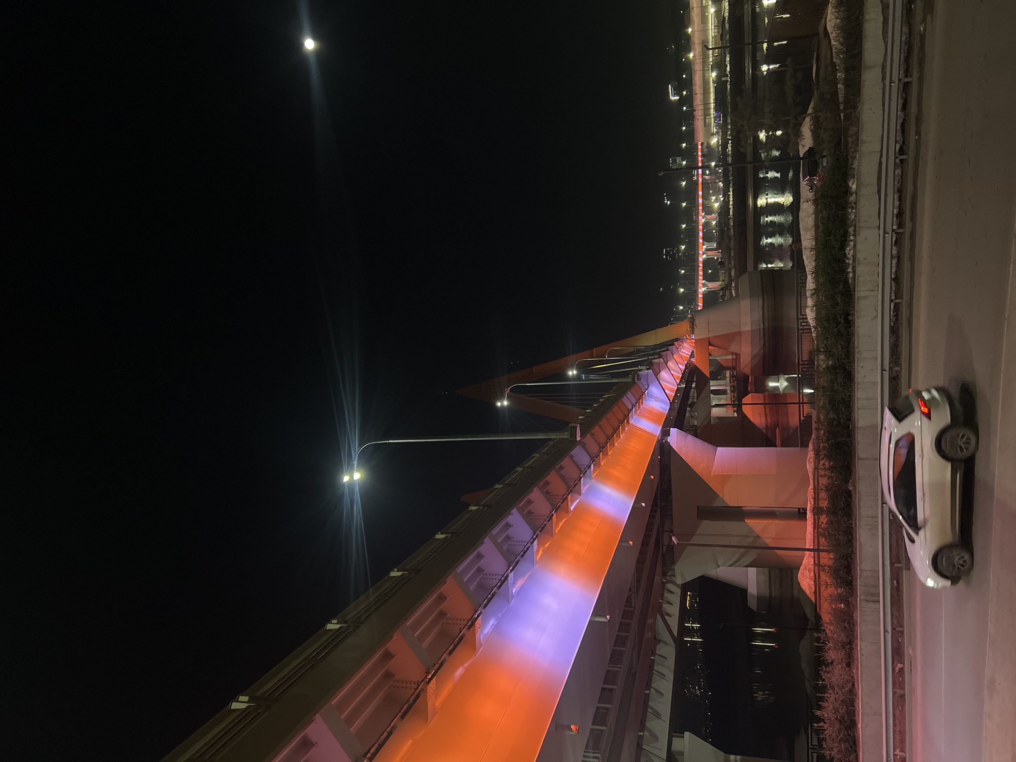 The image captures a night scene in Kazan, Russia, focusing on the illuminated Millennium Bridge. The bridge's structure is prominently displayed, showcasing a vibrant orange and purple color scheme on its underside. A bright moon is visible in the upper left corner of the frame, providing ambient light. A white car is parked on the side of the road running parallel to the bridge. In the background, lights from buildings and reflections on the water contribute to the city's nighttime ambiance. Streetlights also contribute to the illumination of the scene.