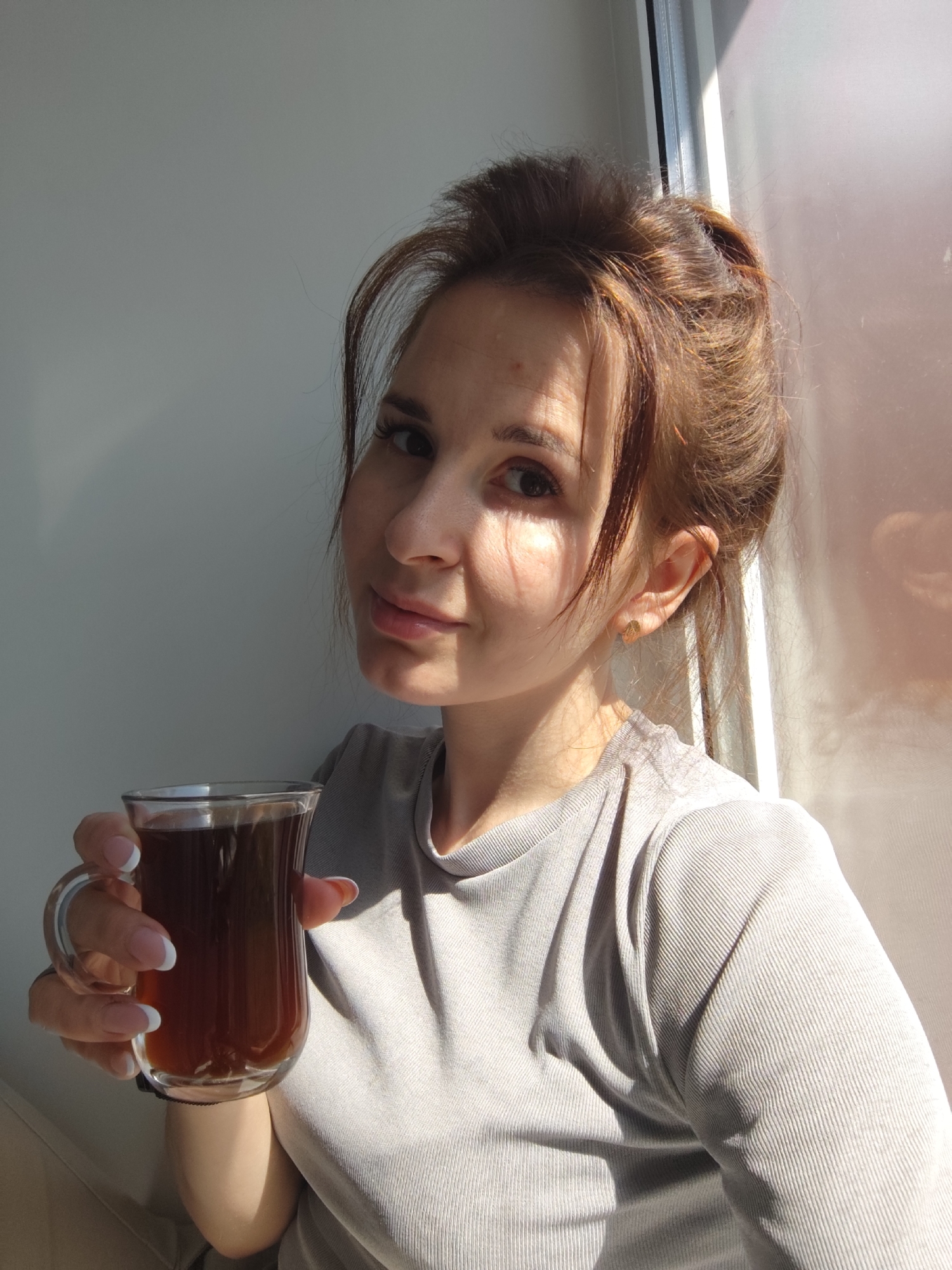 A young woman with light brown hair pulled back into a messy bun holds a glass of dark-colored liquid, possibly tea or coffee, in her left hand. She is wearing a light gray shirt with a ribbed texture. The woman is positioned near a window, with sunlight partially illuminating her face and creating shadows on the wall behind her. Part of the window and curtain is also visible in the background. The overall impression is an informal, possibly at-home setting. There is no evidence to suggest the image was taken in the city of Фоки, Russia.