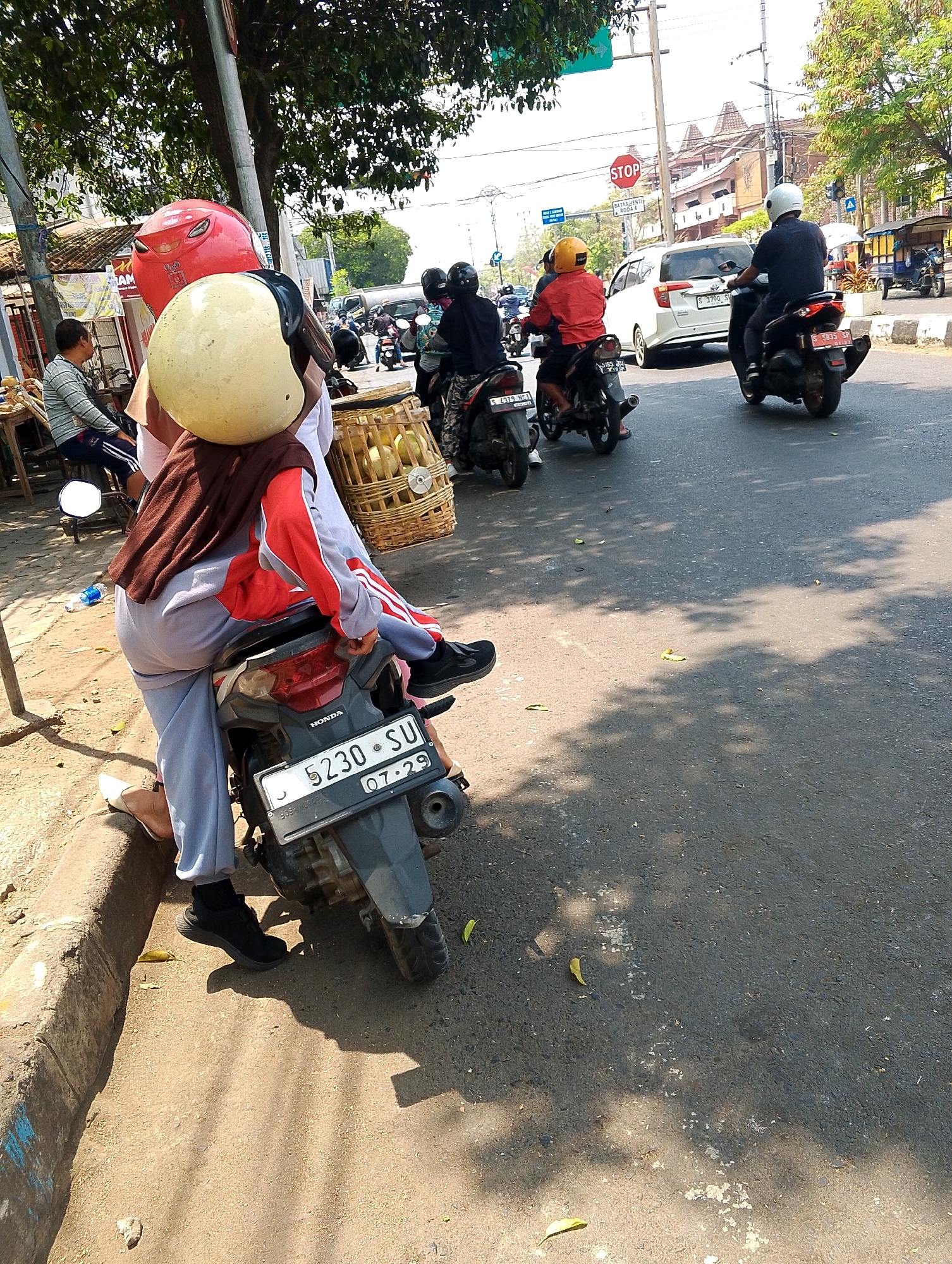 The image depicts a street scene in an urban environment. In the foreground, a person wearing a red and white jacket and gray pants is seated on a black motorcycle. They are wearing a light-colored helmet and a brown headscarf. A basket filled with light-colored, spherical objects is placed behind the rider on the motorcycle. The motorcycle has a license plate that reads "5230 SU 07-29". To the left of the motorcycle, a person wearing a gray and white striped shirt and dark shorts is sitting on a low stool. In the background, other motorcycles and a white car are visible on the road. Traffic signs, including a "STOP" sign, are present. Trees with green foliage line the street, casting shadows on the asphalt. The ground is a mix of dirt and pavement. The weather appears to be sunny.