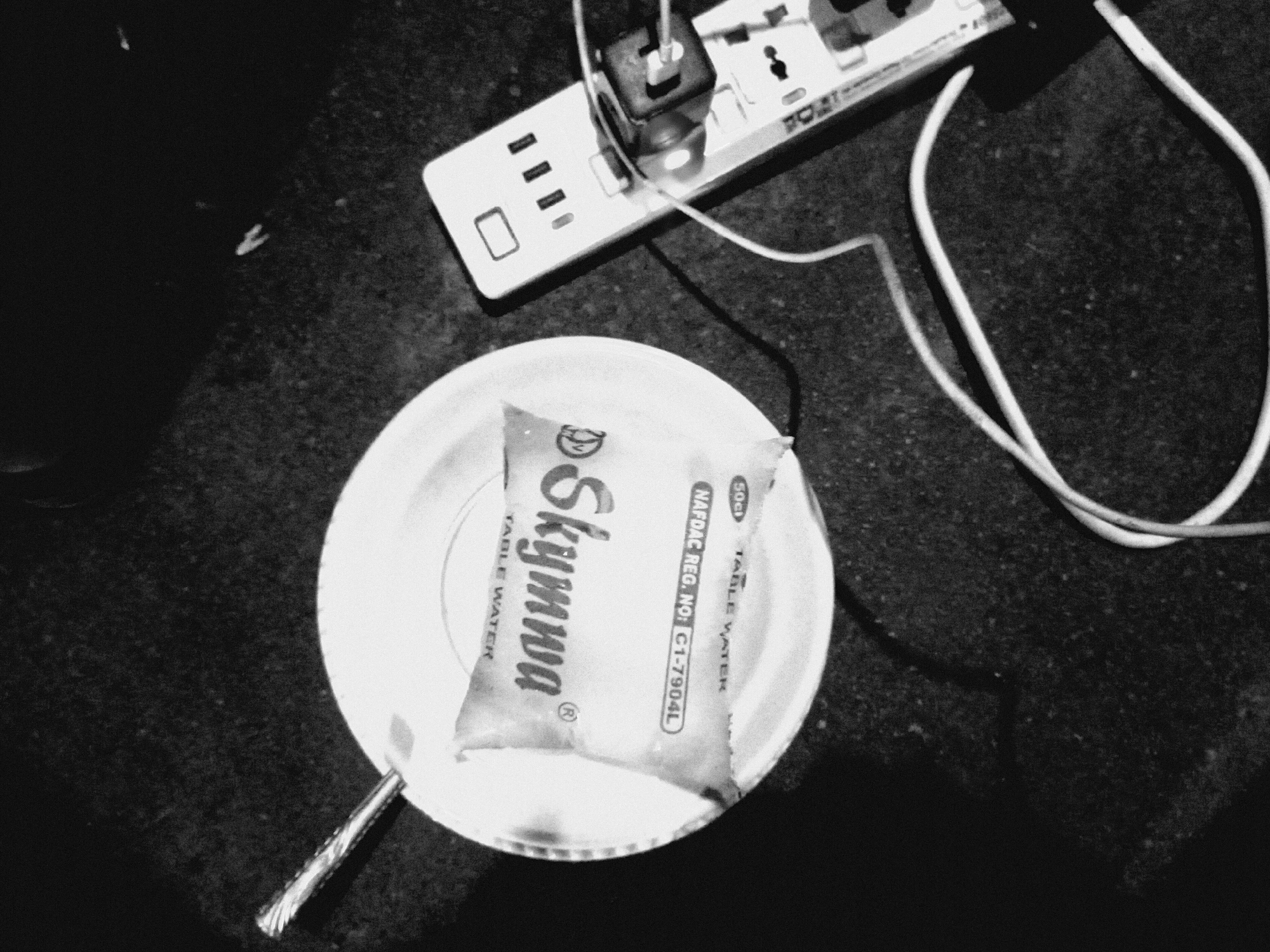 A black and white image depicts a small plastic water bag, labeled "Skymuna TABLE WATER" and "Nafdac Reg. No. C1-7904L", lying on a white plate. A silver spoon rests beside the plate. Above the plate, a white power strip with multiple outlets is visible, with a black adapter plugged into one of the outlets. A white cable extends from the power strip. The scene is set on a dark surface. The location is Unknown, Unknown.