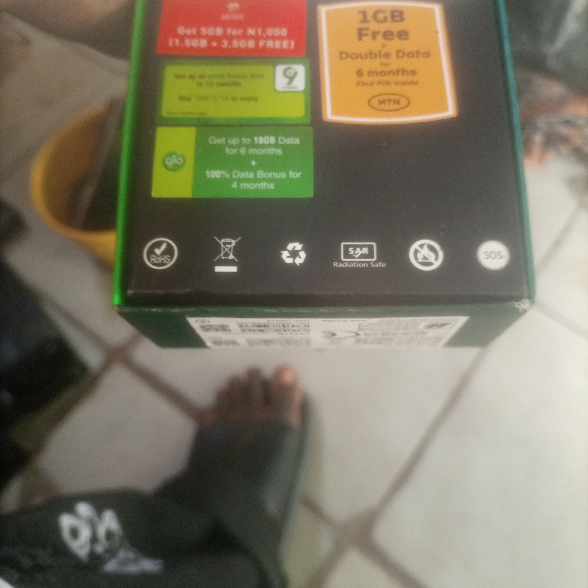 The image shows a close-up of a product box, likely for a mobile phone or data plan, with various promotional offers and certifications printed on it. The box has a green border and features text in red, yellow, and green. Specific offers include "Get 5GB for N1,000 (1.5GB + 3.5GB FREE)," "1GB Free Double Data for 6 months," and "Get up to 18GB Data for 6 months + 100% Data Bonus for 4 months." There are also logos for "glo" and "MTN." Certifications and symbols visible include "RoHS," a recycling symbol, a "SAR Radiation Safe" symbol, a battery disposal symbol, and an "SOS" symbol. The background is blurred, showing what appears to be a tiled floor and a person's foot wearing a dark sock. The setting is indoors, and the lighting suggests a somewhat dim environment. There is no discernible information about the location within a city.