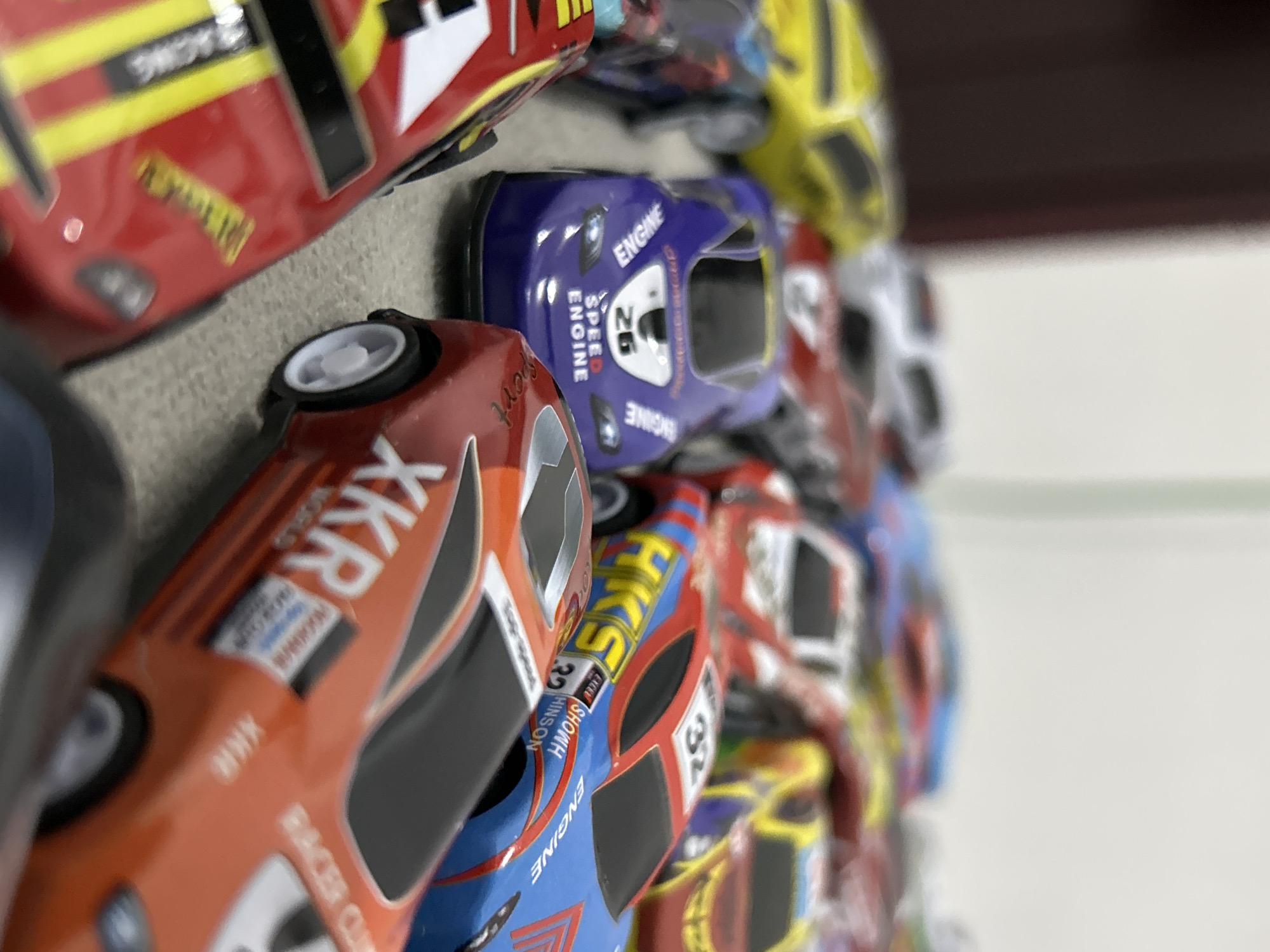 The image displays a close-up, rotated view of a collection of brightly colored toy race cars, likely part of a display or collection, set in Lhokseumawe, Indonesia. The cars are arranged in a line, with several distinct models visible in the foreground and others blurring into the background.
The primary subjects are the miniature race cars, crafted with detailed livery. In the foreground, an orange car is prominent, featuring white lettering and black accents. Behind it, a vibrant blue car stands out with yellow and white details. Further back, red, yellow, and other colored cars are visible, though less distinct due to depth of field. The scene appears to be an indoor setting, with a light-colored surface and a blurred background suggesting a shelf or tabletop against a wall. The lighting is even, implying interior illumination.
Notable details include the racing-themed decals and numbers on the cars. Visible text includes:
- On the orange car: "XKR," "WORLD," "RACER CLUB," and "Sport."
- On the blue cars: "ENGINE" (appearing multiple times), "SPEED," the number "26," "EXCEL SHOWH," "HINSON," "HKS," and the number "32."
- On a red car: "RACING."
The cars appear to be in good condition, showcasing their intricate designs and vibrant aesthetics. There are no people visible, and no specific time of day or weather conditions can be discerned from the indoor setting.