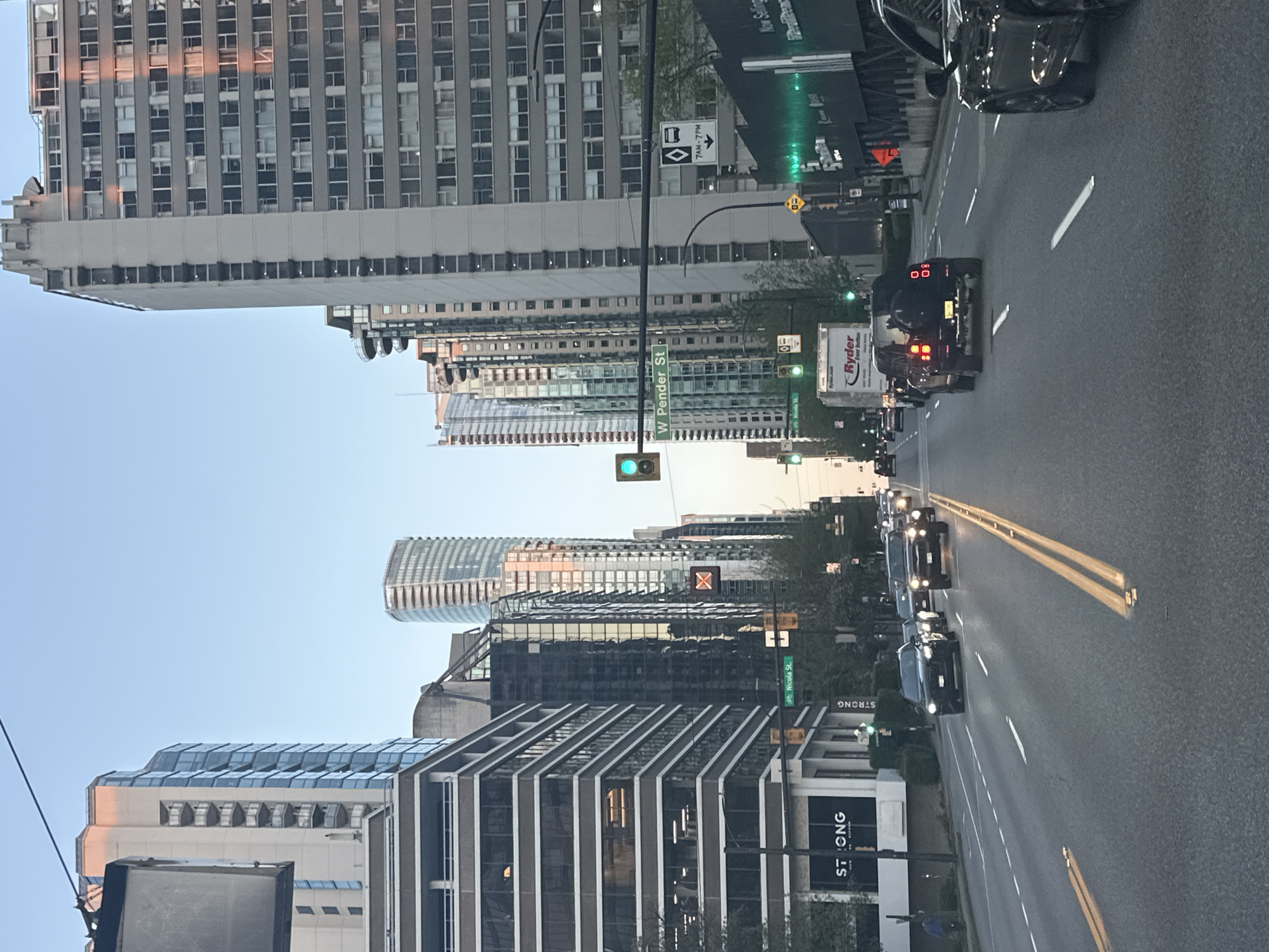 This image captures an upright, eye-level street scene in a bustling urban environment, identified as Vancouver, Canada. The perspective suggests it was taken from a vehicle on a multi-lane road.
The scene is dominated by numerous high-rise and mid-rise buildings lining both sides of the street, showcasing a mix of modern glass architecture with reflective surfaces and older, more traditional structures. The sky above is clear and blue, indicating good weather, with some buildings reflecting the warm hues of what appears to be late afternoon or early evening sunlight.
On the road, several vehicles are visible, including cars and a distinctive white Ryder rental truck. A black SUV or truck is prominent in the foreground, displaying glowing red taillights and what appears to be a digital "00" or similar number on its rear. Traffic appears to be stopped or moving slowly, possibly at an intersection. Green traffic lights are visible, indicating active traffic flow.
Key location and textual details include street signs for "W Pender St" and "Nicola St," confirming the Vancouver context. The white truck bears the "Ryder" logo and the slogan "For Better." Multiple buildings prominently display the word "STRONG." A bus stop sign with a bus icon and an arrow also shows operating hours of "7AM - 7PM." Additionally, a large sign with "Now Selling" is visible on the right. The overall activity depicts a typical, busy city street during daylight hours.