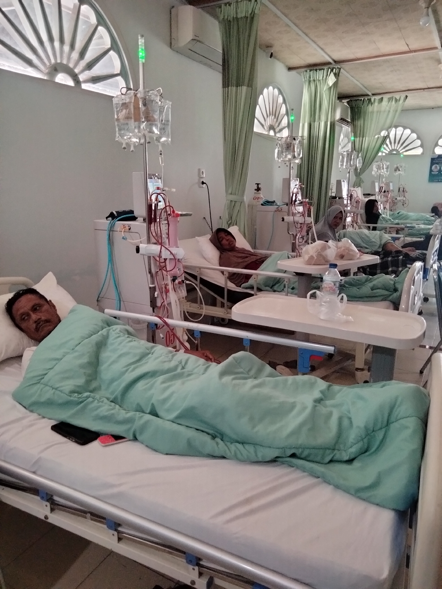 This is a photograph taken inside a room that appears to be a hospital ward, possibly a dialysis center, given the presence of multiple patients connected to medical equipment, including IV drips and dialysis machines. The room is illuminated and features several beds, each occupied by a patient. The architecture includes arched windows with decorative grilles and green curtains that divide the space.
In the foreground, a man lies on a hospital bed covered with a light green blanket. He is looking towards the camera with a serious expression. On the bed beside him, a dark rectangular object, likely a mobile phone, and a pink object are visible.
Further back, other patients are seen in their beds, some with their faces partially obscured. Medical equipment, such as IV stands with multiple bags of fluid, dialysis machines with tubing, and various other medical devices, are positioned around the beds. The overall environment suggests a clinical setting focused on patient care. The presence of arched windows and the specific style of the windows are characteristic of architectural elements found in Lhokseumawe, Indonesia, particularly in older or more traditional buildings within the city.