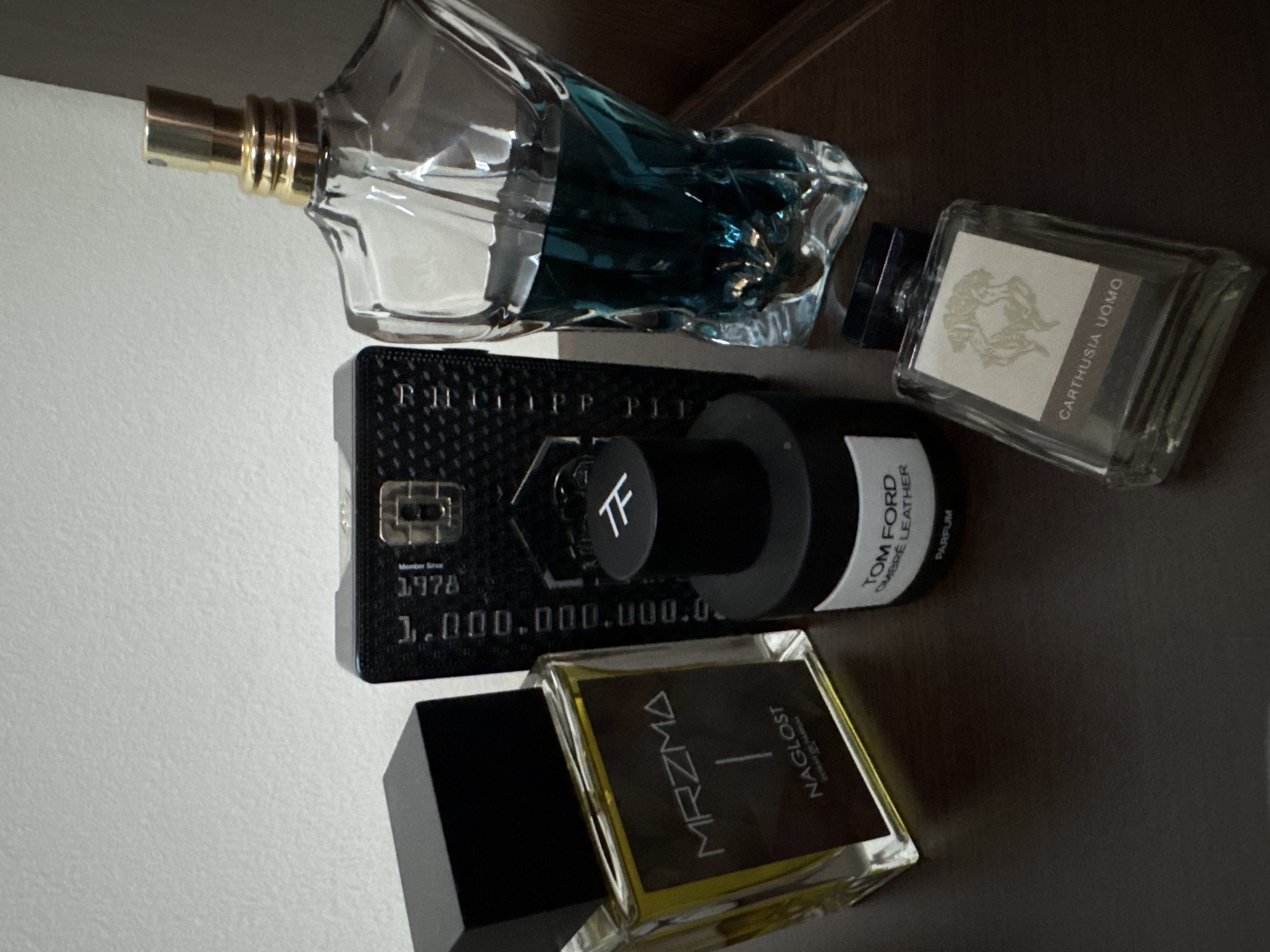 The image shows five bottles of perfume arranged on a surface that transitions from white to brown. The perfumes include:
1. A clear, elaborately shaped bottle with a gold-colored nozzle. The perfume inside is a dark blue color.
2. A black rectangular bottle with a textured surface, bearing the name "PHILIPP PLEIN". It has a cylindrical atomizer labeled with "TF". The text "Member Since 1978" and "1,000,000,000,000" is visible on the bottle.
3. A cylindrical bottle labeled "TOM FORD OMBRE LEATHER PARFUM" with a black cap.
4. A clear rectangular bottle with a label indicating "CARTHUSIA UOMO".
5. A clear rectangular bottle with a black cap and a label that reads "MRZMA NAGLOST EXTRAIT DE PARFUM".
The location, Sofia, Bulgaria, is not represented in the image.