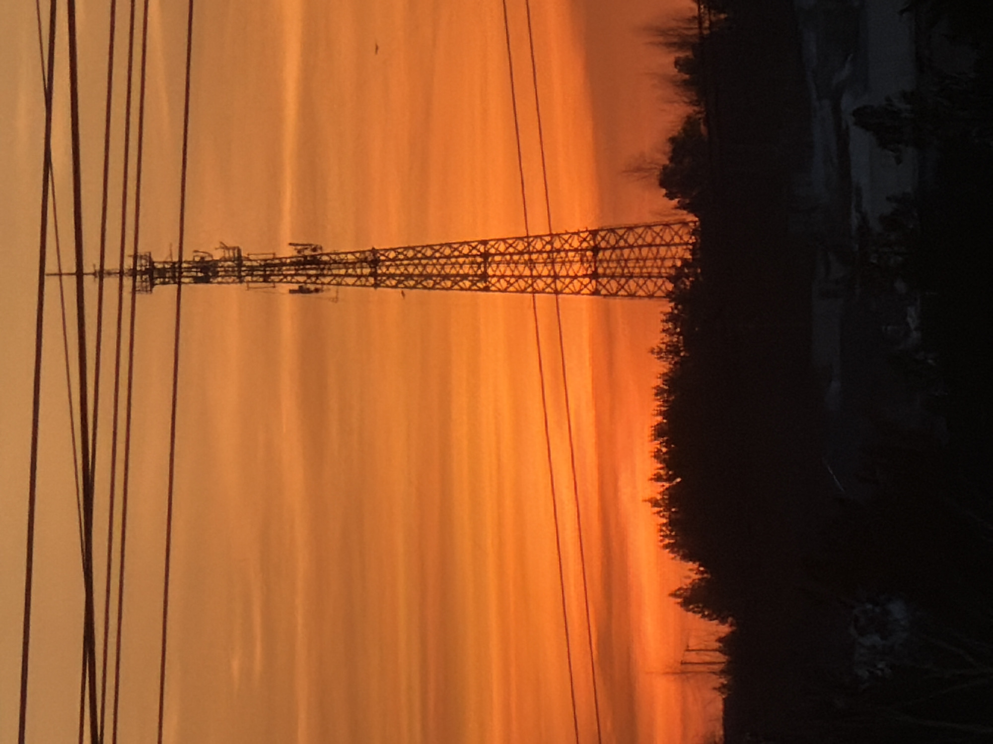 Here is a concise description of the image:
The image depicts a transmission tower against a vibrant orange and yellow sunset sky. The tower is silhouetted and stands tall, with a lattice structure. To the right of the tower is a dark treeline silhouetted. Electrical power lines run vertically in the foreground. The location is potentially Chaikovsky, Russia, as stated.