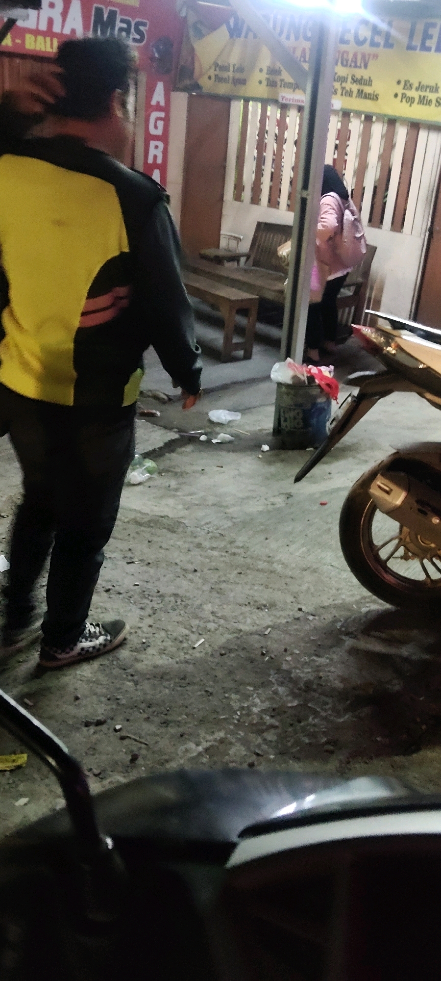 A vertical, medium shot captures a scene outside a warung (small shop) in Sukaresmi, Indonesia. A person wearing a black and yellow jacket and black pants stands in the foreground, facing away from the camera. Their right hand is visible, held near their waist. In the mid-ground, to the right of the first person, a second person wearing a pink jacket and black pants walks away from the camera, carrying a pink bag over their shoulder. The warung has a red and yellow sign that reads "WARUNG PECEL LELE" and "AGRA Mas." Wooden benches are placed in front of the warung. A blue and white trash bin filled with red and white plastic bags is situated in front of the warung. The ground is concrete and appears dirty, with scattered cigarette butts and plastic waste. A portion of a motorcycle is visible in the lower right corner of the frame. The lighting suggests it is evening or nighttime, with artificial light illuminating the area.