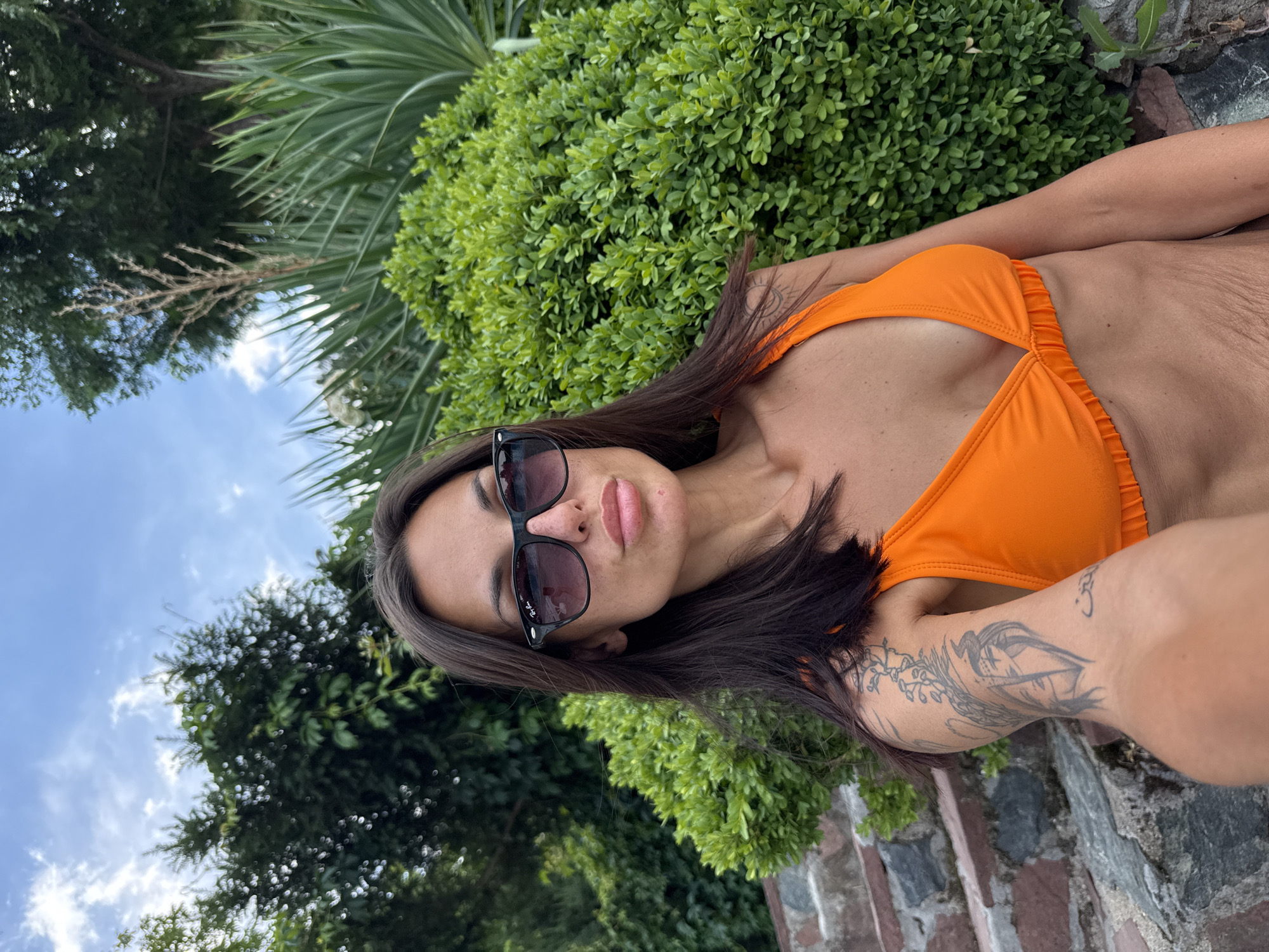 A woman with dark hair, sunglasses, and an orange bikini top poses for a selfie outdoors. She is leaning against a stone wall and is surrounded by lush green bushes and trees, with a cloudy blue sky overhead. Her left arm is visible, showing tattoos. The location is in the city of Grivitsa, Bulgaria.
