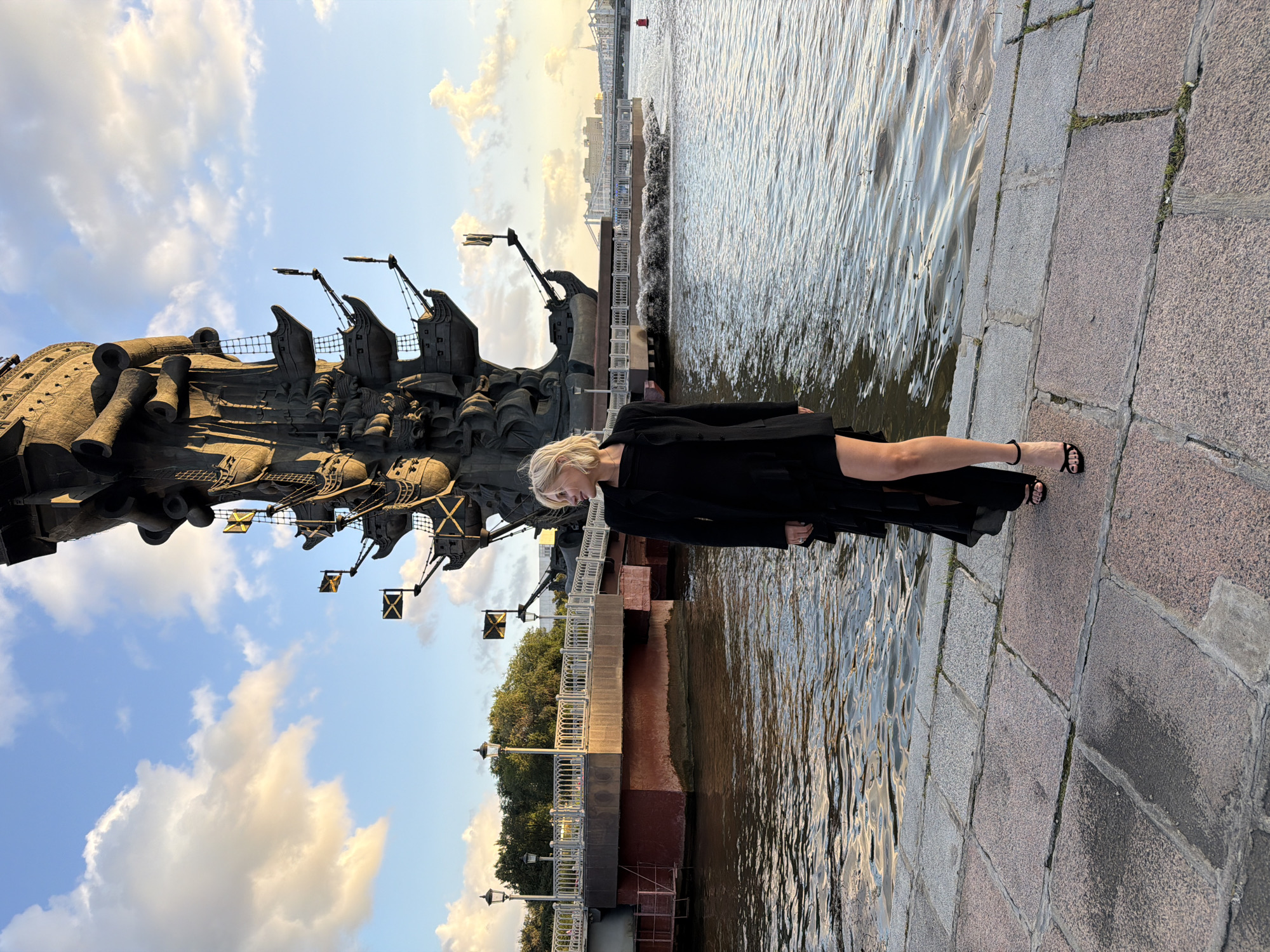 A blonde woman wearing a black dress and black high heels stands on a stone embankment overlooking a body of water. Behind her is a large, dark sculpture of a ship with multiple levels and cannons. The sculpture is situated near a bridge with decorative lampposts. The sky is blue with scattered white and yellow clouds, suggesting it is late afternoon or early evening. In the distance, more bridges and buildings are visible, indicating an urban setting. The sculpture is identified as the Peter the Great Statue, a prominent landmark on the Moskva River in Moscow, Russia.