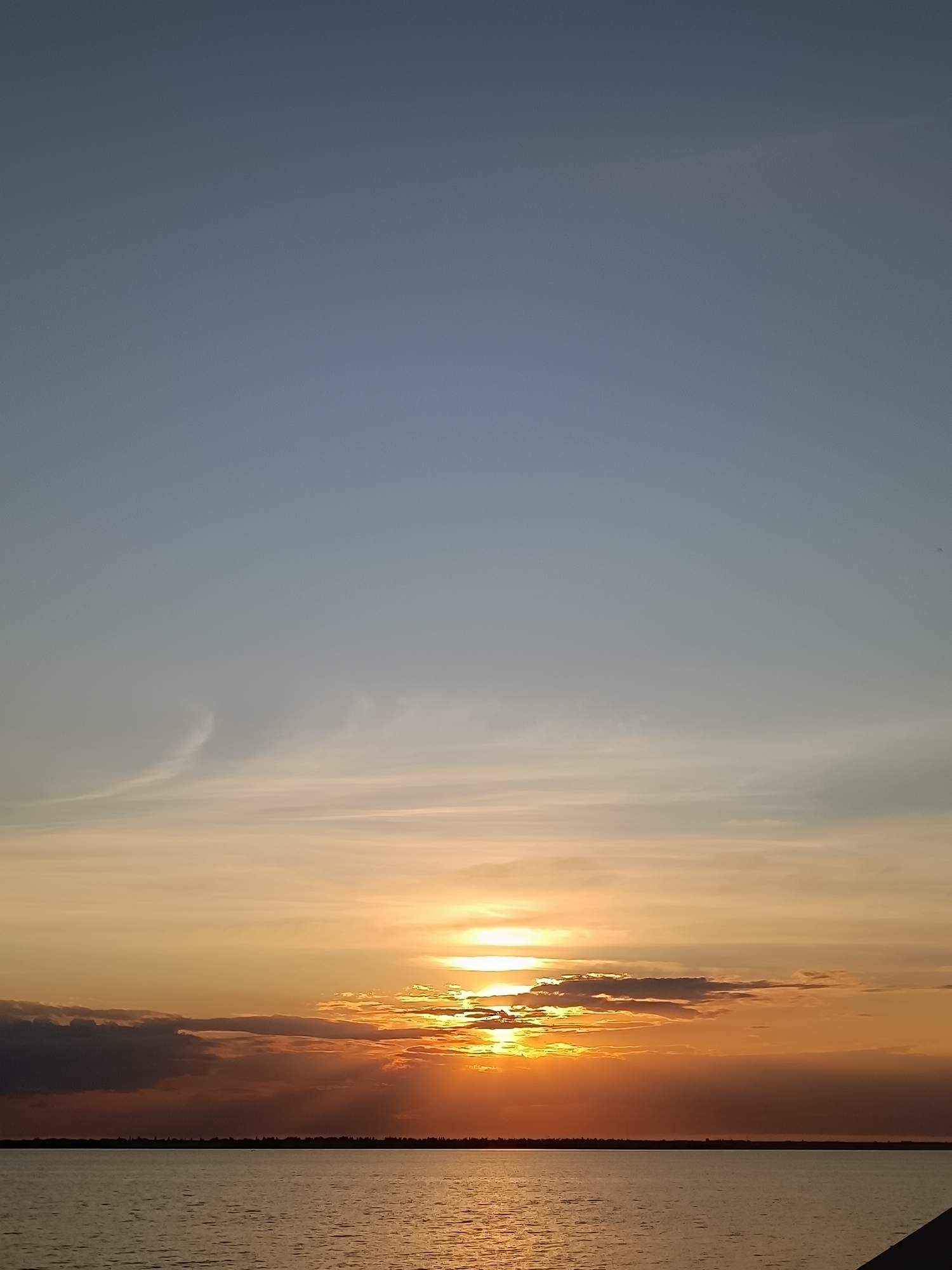 An aerial shot captures a serene sunset over a body of water, likely a lake or ocean, extending towards a distant horizon. The sky transitions from a pale blue at the top to a warm orange and yellow as it nears the horizon. Wispy clouds are scattered across the sky, with denser, darker clouds partially obscuring the setting sun, which casts a golden glow on the water's surface. The far horizon is lined with a dark, silhouetted landscape, suggesting a city or a dense forest. The water is calm, with gentle ripples reflecting the vibrant colors of the sky. The perspective is from a slightly elevated position, looking out over the vast expanse of water and sky.