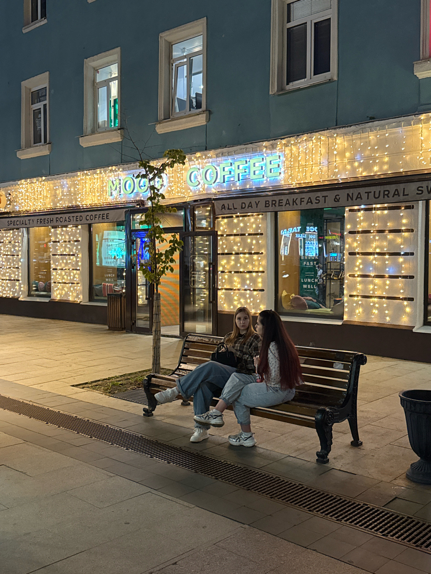 The image depicts a street scene at night in Tver, Russia, in front of a brightly lit cafe. Two young women are seated on a park bench. The cafe facade is decorated with strings of warm white lights, and neon signs are visible through the windows. One sign reads "MOON COFFEE" in green neon. Another sign above the entrance states "SPECIALTY FRESH ROASTED COFFEE," and a banner below the windows advertises "ALL DAY BREAKFAST & NATURAL SW." The women are casually dressed, with one wearing a plaid shirt and jeans, and the other a patterned blouse and jeans. They appear to be talking to each other, with the woman in the patterned blouse holding a drink. The sidewalk is paved with stone tiles, and a metal grate runs along the edge of the walkway. A dark trash receptacle is visible to the right of the bench. The building behind the cafe is a multi-story structure with windows. The overall atmosphere is that of a casual evening gathering outside a cafe.