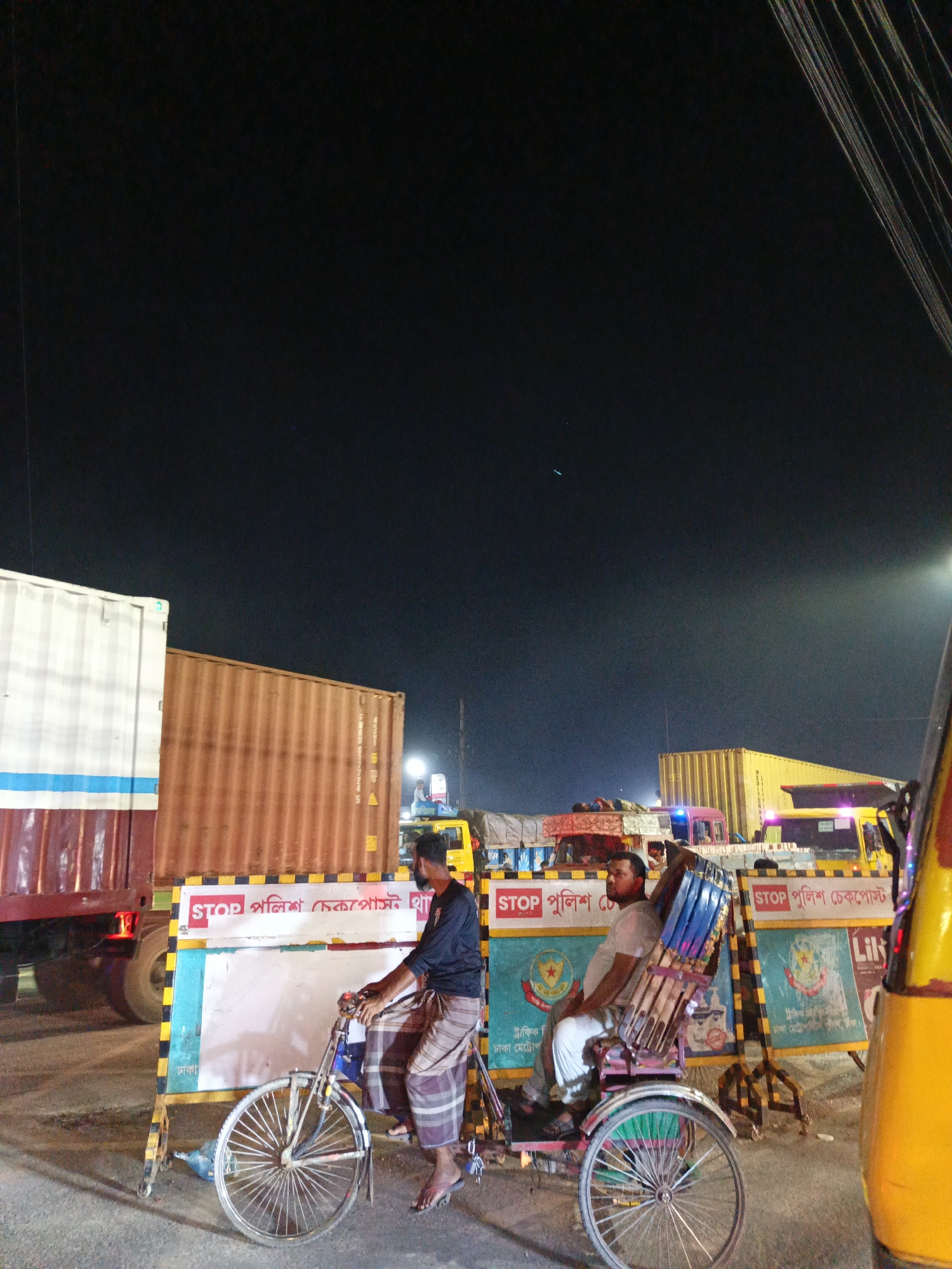 Here is a detailed description of the image:
**Scene:** An outdoor scene at night, likely a checkpoint or traffic stop area in an urban environment. The image is dominated by large trucks and barriers, along with people on a cycle rickshaw.
**People:**
* A man is riding a cycle rickshaw. He wears a dark long-sleeved shirt and a checked lungi (a type of sarong).
* A passenger sits in the back of the rickshaw. He is wearing a light-colored shirt and white trousers.
**Objects:**
* A cycle rickshaw with a blue and purple seat cushion.
* Several large cargo trucks are in the background, some with containers.
* Barriers with signs are placed along the road. The signs have the words "STOP পুলিশ চেকপোস্ট" (STOP Police Checkpost) written in Bengali.
**Interactions:**
* The rickshaw driver is actively pedaling his vehicle.
* The passenger is sitting on the rickshaw.
**Environment:**
* The scene appears to be lit artificially.
* The sky is dark, indicating it is nighttime.
* Electrical wires are visible in the top right of the frame.
* A yellow autorickshaw is visible on the right side of the image.
**Location:**
* The text on the sign suggests that the scene is in Dhaka, the capital city of Bangladesh.