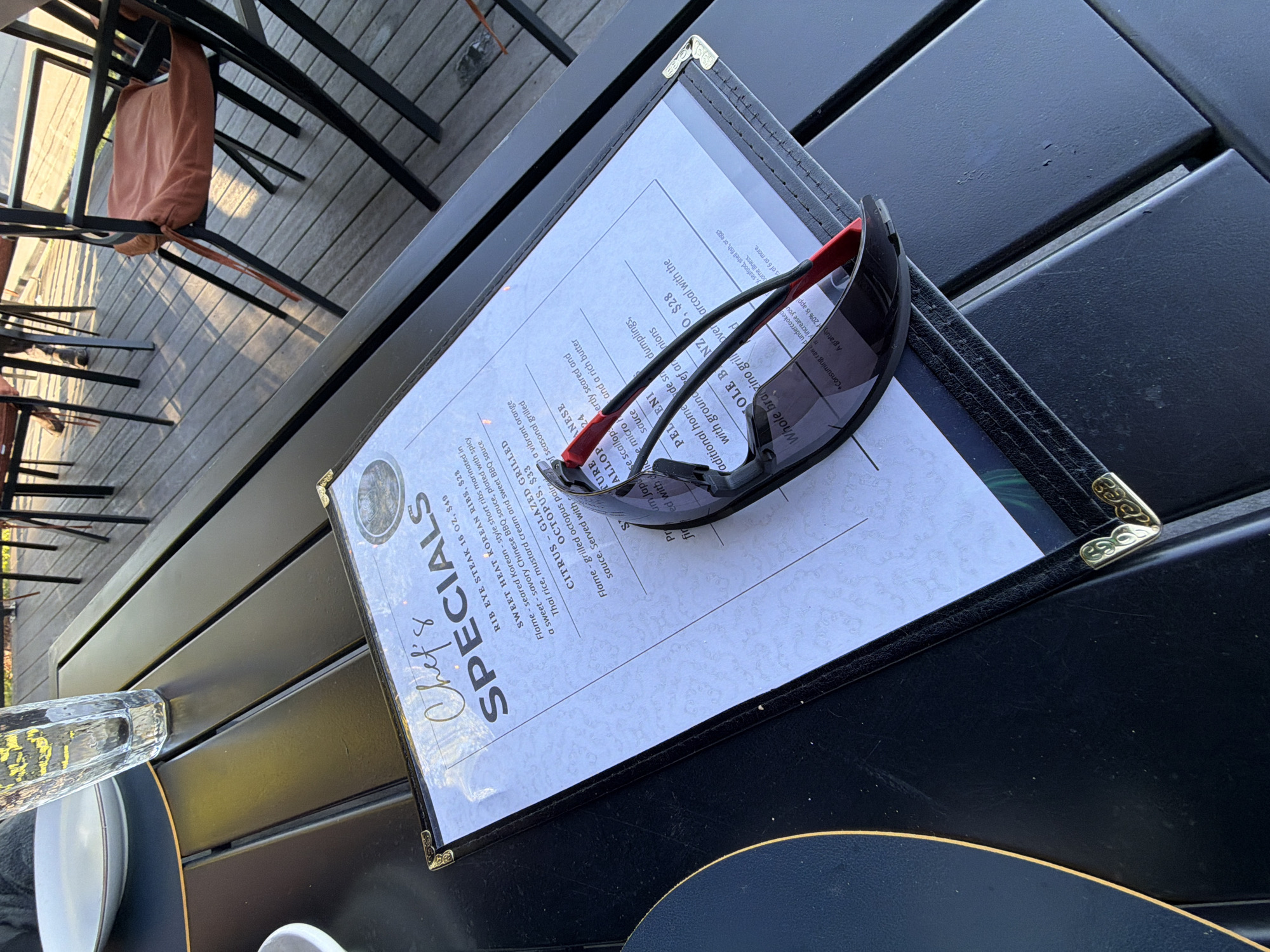 A black outdoor table, likely at a restaurant in Glenview, United States, holds a menu featuring "Chef's Specials." A pair of sunglasses rests on the menu. Tableware is visible, suggesting a meal setting. The table itself is made of dark planks, and chairs with fabric cushions are partially visible, indicating an outdoor dining area. The scene implies someone is about to dine or has just finished a meal.
