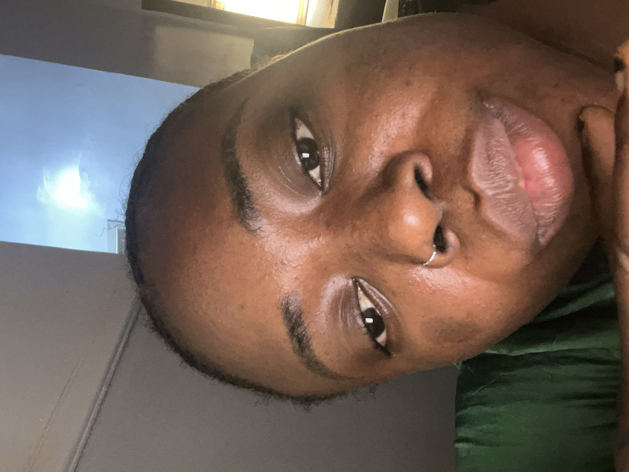 A close-up, angled shot of a dark-skinned person with a nose ring. The person is wearing a dark green garment. The background shows a white wall and what appears to be a window with light coming through it. The location in Kubwa, Nigeria, is not evident from the image.