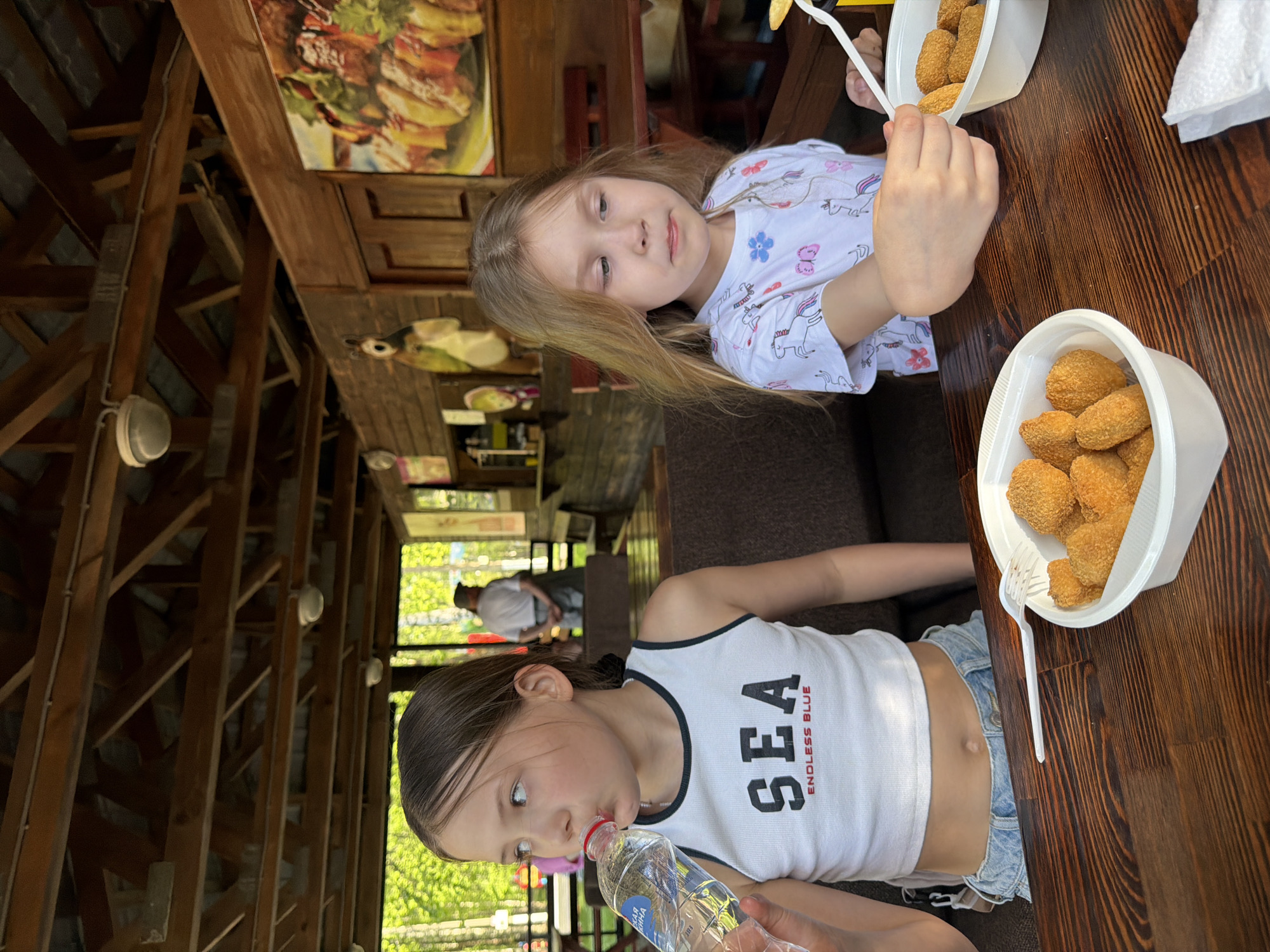 Two young girls are seated at a wooden table in an outdoor eating area. The girls are eating from two white plastic containers filled with golden-brown, nugget-like food. One girl is drinking from a plastic water bottle. The outdoor eating area has a wooden frame structure. A man is visible in the background, outside the structure. A sign depicting a burger is affixed to an interior wall. The location is Chaikovsky, Russia.