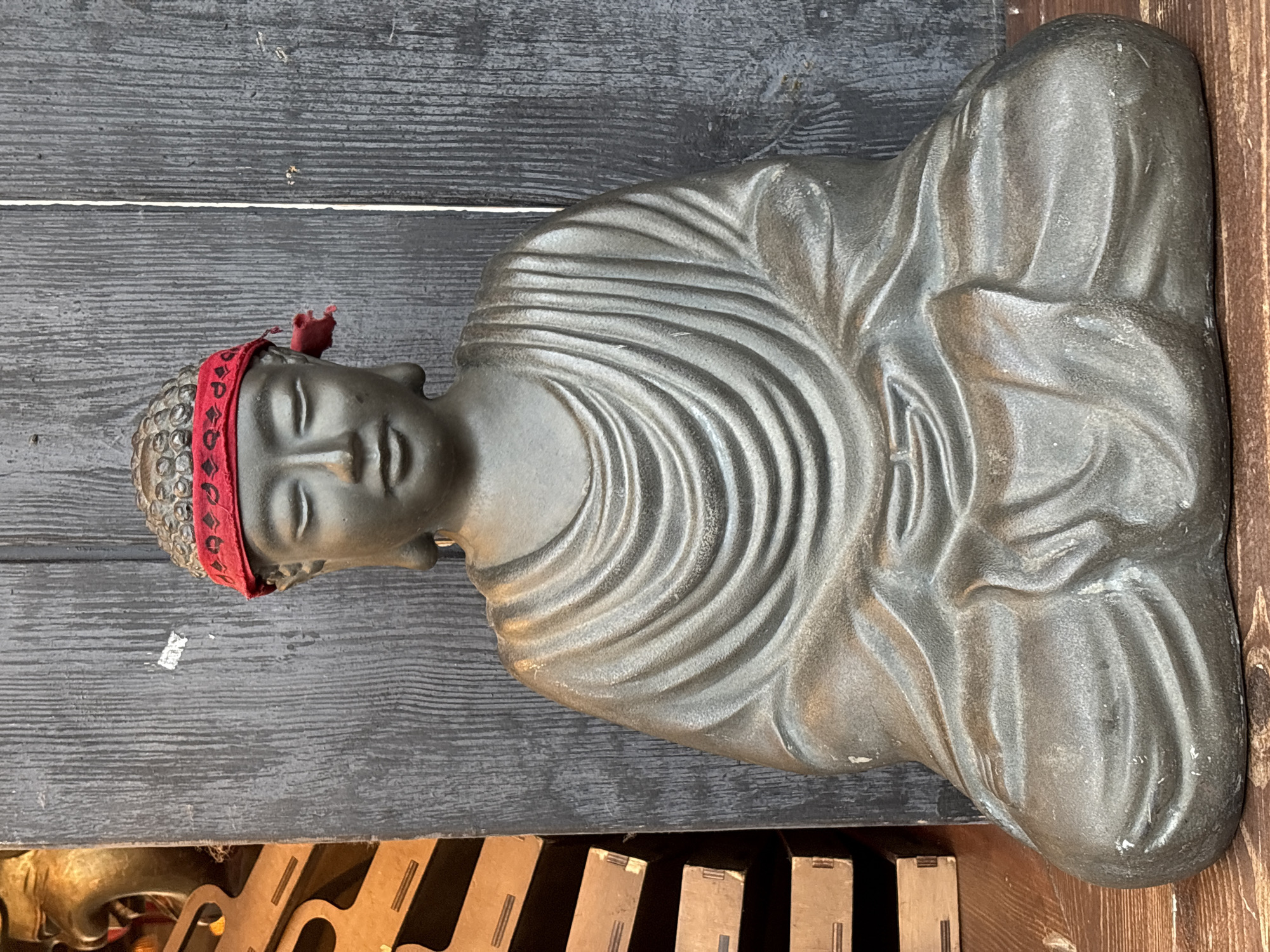 A dark, weathered statue of a meditating Buddha is depicted, rotated 90 degrees clockwise in the image. The Buddha is sculpted with a serene expression, eyes closed, and its head features small, rounded bumps, possibly indicating hair. A striking red fabric headband with a dark pattern is tied around its forehead, with a small, frayed piece extending upwards from the crown. The statue's body is covered in flowing robes, rendered with distinct, deep folds and creases, suggesting a heavy material like metal or stone with a slightly textured surface and subtle highlights.
The background behind the statue is a dark, horizontally planked wooden wall or surface, showing some wear and tear, including minor white scuffs and streaks. A thin, horizontal white line is visible across the wooden background, running behind the Buddha's head. In the lower portion of the image, below the Buddha's base, are indistinct objects, possibly wooden or metallic structures, with geometric shapes and varied tones, suggesting parts of furniture or another architectural element. The overall lighting appears soft and even, highlighting the textures of the statue and its surroundings. There is no visible text. The location context is Kyiv, Ukraine.