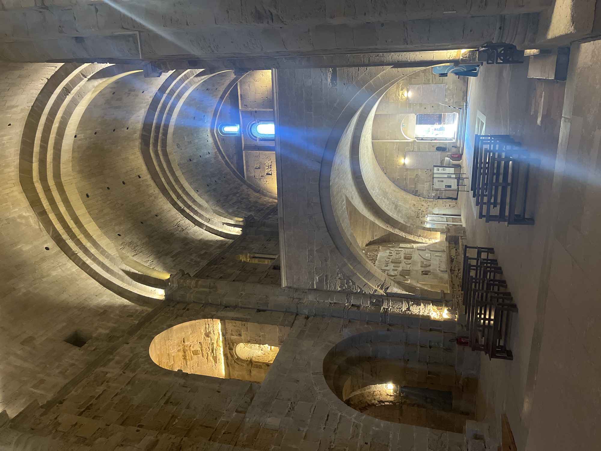 This is an interior view of the Cathedral of Saint-Pierre-et-Saint-Paul in Villeneuve-lès-Maguelone, France. The architecture features large stone arches supporting a vaulted ceiling. Natural light streams through high windows, casting bright shafts of blue and white light across the stone floor and illuminating the dusty atmosphere. In the foreground, rows of dark wooden benches are arranged, suggesting a seating area for visitors or worshippers. The walls are constructed of rough-hewn stone blocks, with visible architectural details and some areas illuminated by artificial lights, giving a warm glow. In the distance, an open doorway allows a view of bright sunlight and the exterior. A few informational panels and fire extinguishers are visible along the walls. The scene is devoid of any apparent activity or human interaction, conveying a sense of quiet and historical solemnity.