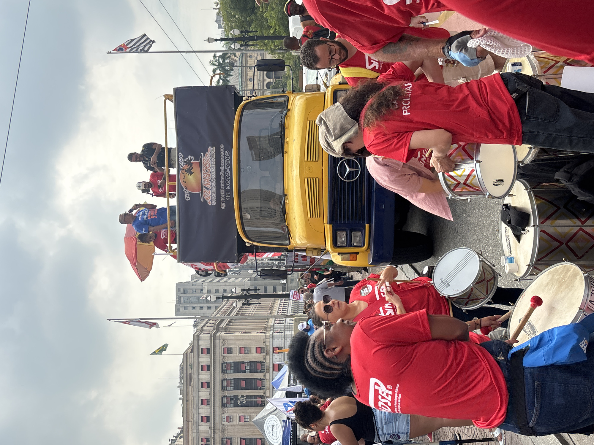 The image captures a vibrant scene in São Paulo, Brazil, featuring a political demonstration or parade.
Foreground: A group of people wearing matching red t-shirts are playing drums. Some are actively drumming, while others are resting. The drums vary in size and color, and they are placed close together on the ground.
Midground: Behind the drummers, there's a large yellow truck with a blue front. A sign on the truck reads, "TRIO ELETRIDO SOL NASCENTE," along with a phone number. People are visible on the top of the truck. In the background are buildings with classical architectural designs, complete with columns and numerous windows. Flags are waving in the distance, including one of São Paulo, and one of Brazil.
Background: The sky is overcast, suggesting it might be a cloudy day. There are other people in the background, participating in the event.
Overall: The atmosphere suggests a lively, energetic gathering, likely a political rally or cultural celebration.