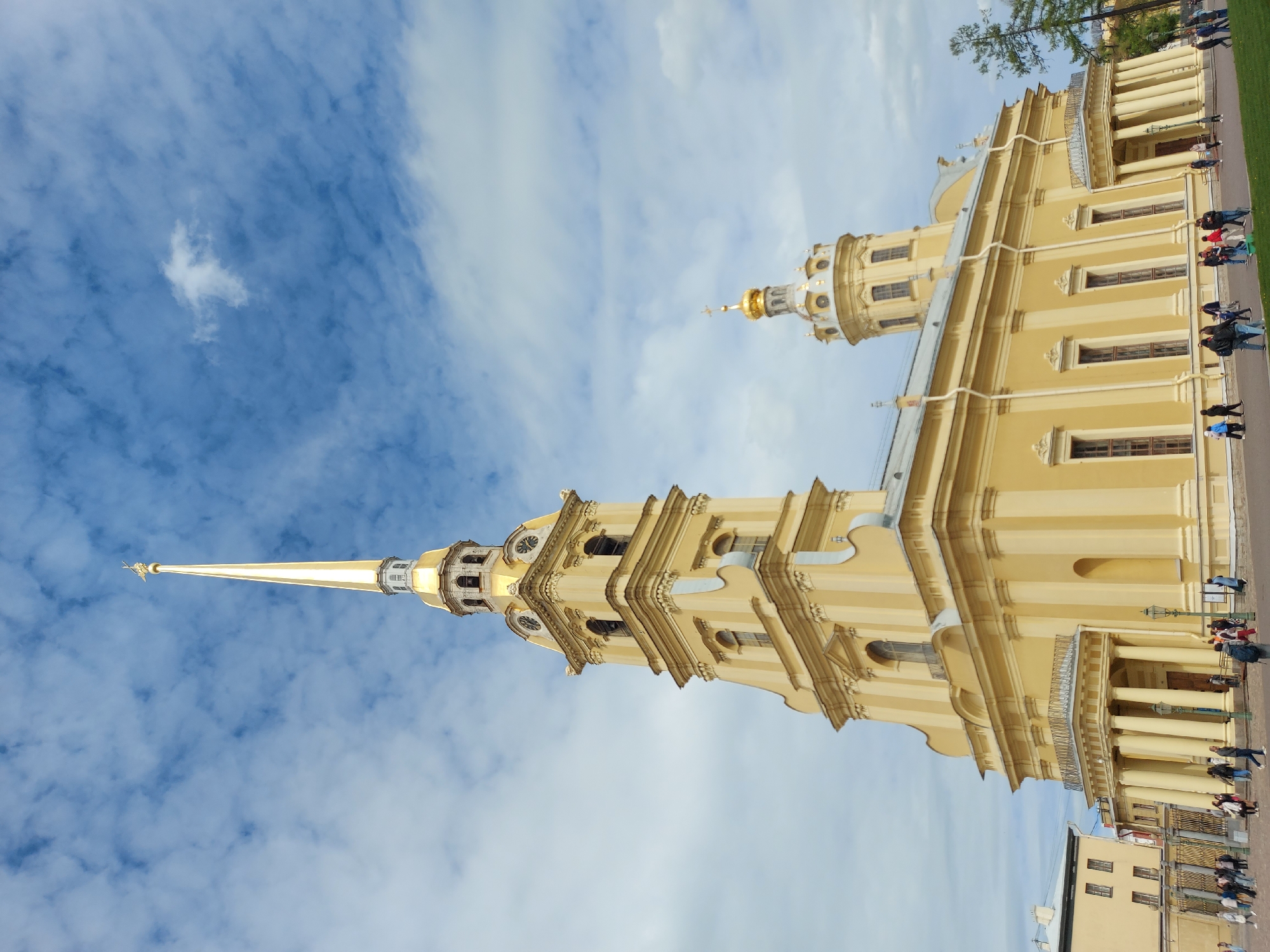 The image is a vertical shot of the Peter and Paul Cathedral in Saint Petersburg, Russia. The cathedral is a tall, yellow baroque-style building with a prominent spire topped by a golden angel. The building features columns, windows, and detailed architectural elements. The sky is blue with scattered white clouds. People are walking on the ground near the building. The photo is taken from a low angle, looking up at the cathedral against the sky.