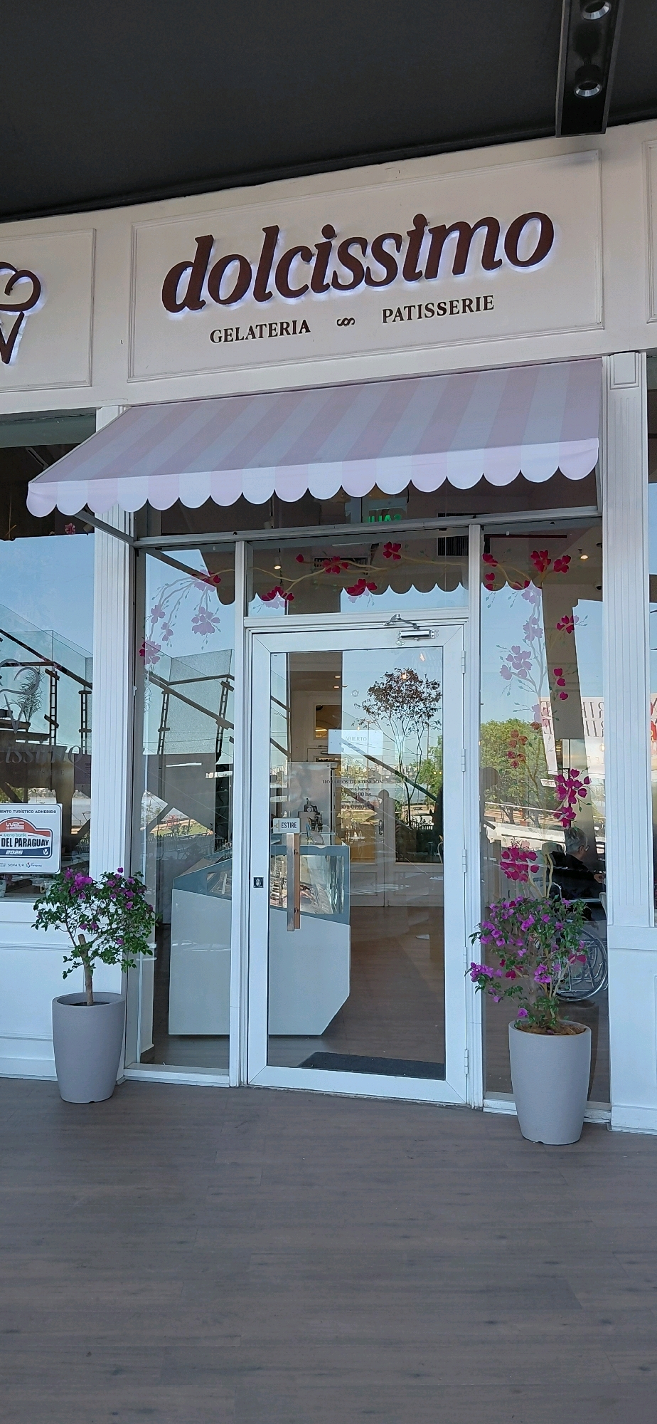 The image shows the storefront of "dolcissimo", a gelateria and patisserie in Encarnación, Paraguay. The facade features a white awning with pink stripes and scalloped edges. Large glass windows display the interior, where refrigerated display cases for gelato and pastries are visible. A glass door with silver handles leads into the shop. Two potted plants, one with green leaves and another with pink flowers, flank the entrance. A sign on the left window indicates the store is part of "WBC Paraguay 2026". The flooring in front of the shop is a dark wood laminate. Reflections in the glass show trees and the sky, as well as a person in a wheelchair inside the establishment. The overall ambiance is clean and modern.
