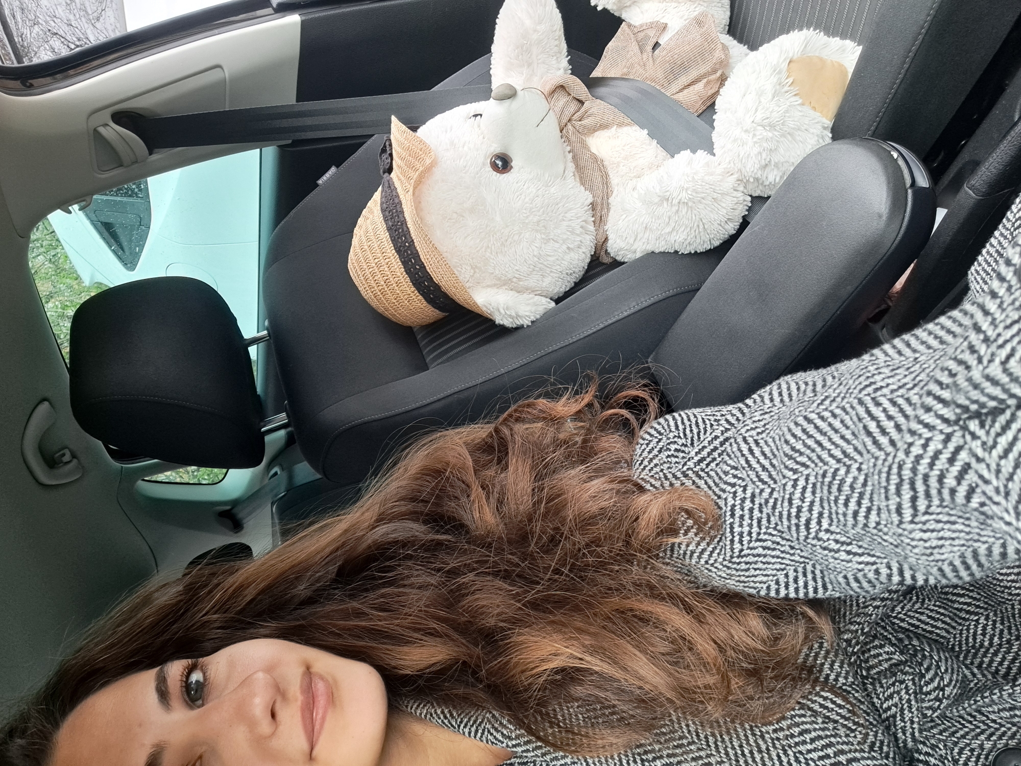 A selfie-style photograph taken inside a car, likely in Sofia, Bulgaria, given the context provided. The angle is from the passenger seat, looking slightly downwards. A young woman with long, wavy, brown hair is visible in the foreground, wearing a herringbone patterned grey and white coat or jacket. She is smiling subtly.
The passenger seat next to her is occupied by a large, white, stuffed bunny wearing a straw hat with a dark band and a light brown scarf. The bunny is secured with a seatbelt.
The interior of the car is primarily black and grey. Outside the passenger-side window, a glimpse of a white vehicle and some greenery is visible, suggesting an outdoor setting. The car's interior ceiling is partially visible, revealing a sunroof.