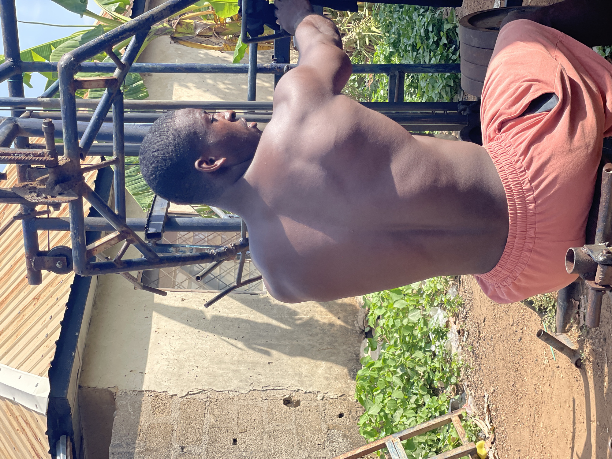 A shirtless man is hanging from a metal exercise machine in Akwanga, Nigeria. He is wearing pink shorts. The machine appears to be outdoors, with a wall, foliage, and a dirt ground visible behind him. The exercise machine looks old and partly rusted. There are weight plates on one end.