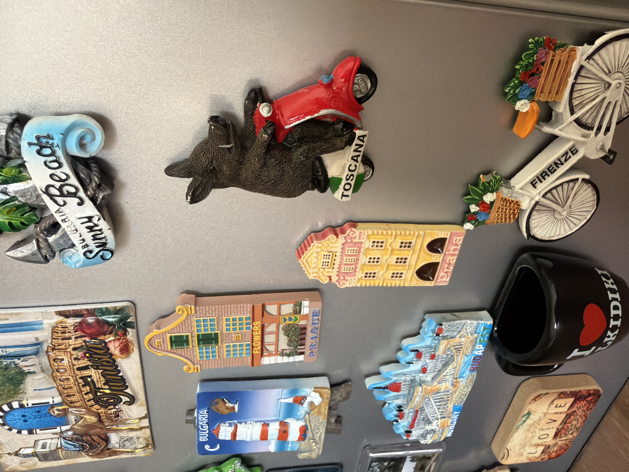 The image displays a collection of refrigerator magnets. From top to bottom and left to right, the magnets show: a scene of Tunisia with a camel and ruins; a magnet with text "Sunny Beach Bulgaria"; a magnet depicting a lighthouse in Bulgaria; a Dutch-style house magnet labeled "Flowers Prague"; a magnet of a wild boar riding a red scooter labeled "Toscana"; a magnet of a multi-story building labeled "Praha"; a magnet of a bridge labeled "Hungary Budapest"; a rectangular magnet with the word "LOVE"; a black mug magnet with a red heart and the text "I KIDIKI"; and a white bicycle magnet with flower baskets labeled "FIRENZE". The location in the city of Pazardzhik, Bulgaria is not depicted in this image.