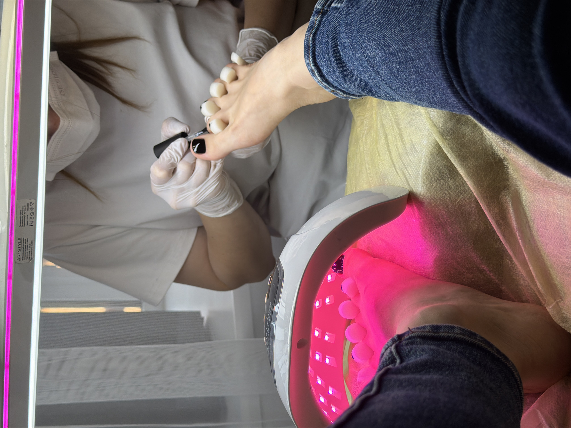This close-up, top-down view captures a pedicure session in what appears to be a nail salon in Perm, Russia. The image is oriented vertically.
In the lower right, a client's foot, partially covered by blue denim jeans, is resting inside a white UV/LED nail lamp which emits a bright pink/purple light, indicating a gel polish curing process. Toe separators are visible on the toes inside the lamp.
To the upper left, a pedicurist, wearing a white uniform, white disposable gloves, and a white face mask, is diligently applying black nail polish with a distinct white design to the big toenail of the client's other foot. This foot also has white toe separators in place. The pedicurist's brown hair is visible behind her mask.
A light yellow or beige textured fabric is draped across the middle of the frame, underneath the client's leg. On the far left, a modern-looking vertical light fixture with a prominent pink strip and a white strip illuminates the scene. A label on this fixture is partially visible, showing text such as "ARTSTYLE" and "EAC". The overall lighting suggests an indoor, professional setting. The activity is clearly a detailed foot care service, likely involving gel nail polish application.