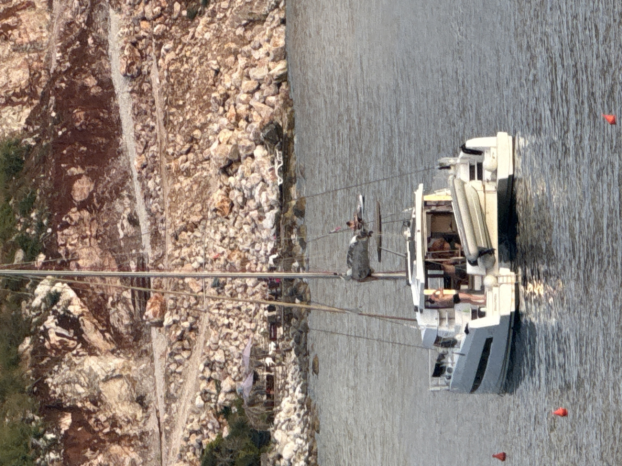 In Kamenovo, Montenegro, a white catamaran is anchored in calm water near a rocky, excavated hillside. The catamaran has a gray inflatable dinghy secured to its stern. On the deck of the catamaran, a person in black shorts stands near the railing. A beach area with two thatched umbrellas and loungers is visible on the shore. The sea is a muted gray-blue, reflecting the overcast sky. Red buoys are scattered in the water.