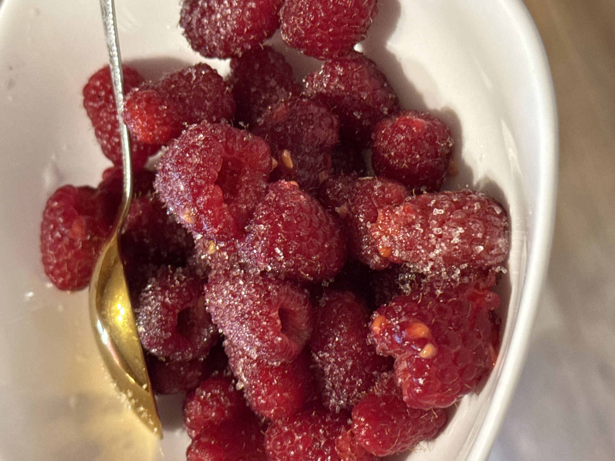 A close-up, overhead shot captures a white bowl overflowing with fresh raspberries. The berries are coated in a shimmering layer of sugar, giving them a frosted appearance. A gold spoon rests partially submerged in the raspberries, its polished surface reflecting the light. Another metal utensil, possibly a fork, is also visible, nudging some of the berries. The background is a blurry expanse of soft light, suggesting an indoor setting, possibly a kitchen or dining area.
