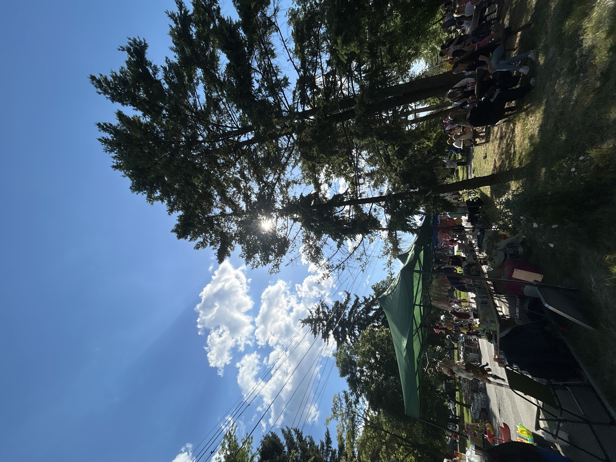 The image shows an outdoor event in Mirovyane, Bulgaria. A row of people is seated on a grassy area in the upper-right. Trees cast shadows across the grass. Below the trees, a green tent covers a line of tables with merchandise, likely part of a market or fair. Underneath the tent, objects and items are displayed for sale. In the foreground, chairs and other market equipment are visible. Above, the sky is blue with scattered clouds, with sunlight streaming through the trees. Power lines run across the sky.