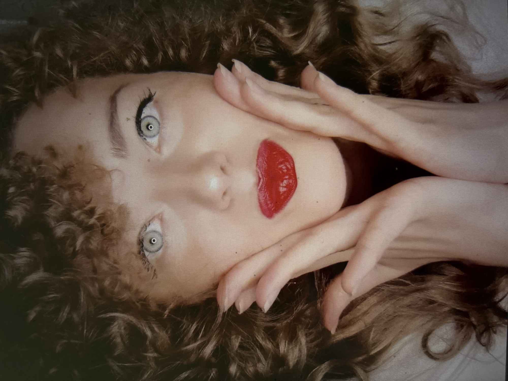 The image shows a close-up portrait of a woman with curly brown hair and light blue eyes. Her face is framed by her hair, and her hands are placed on her cheeks. She is wearing bright red lipstick. The background is dark and out of focus. There is no indication of the city of Novosibirsk, Russia, or any other specific location within the image.