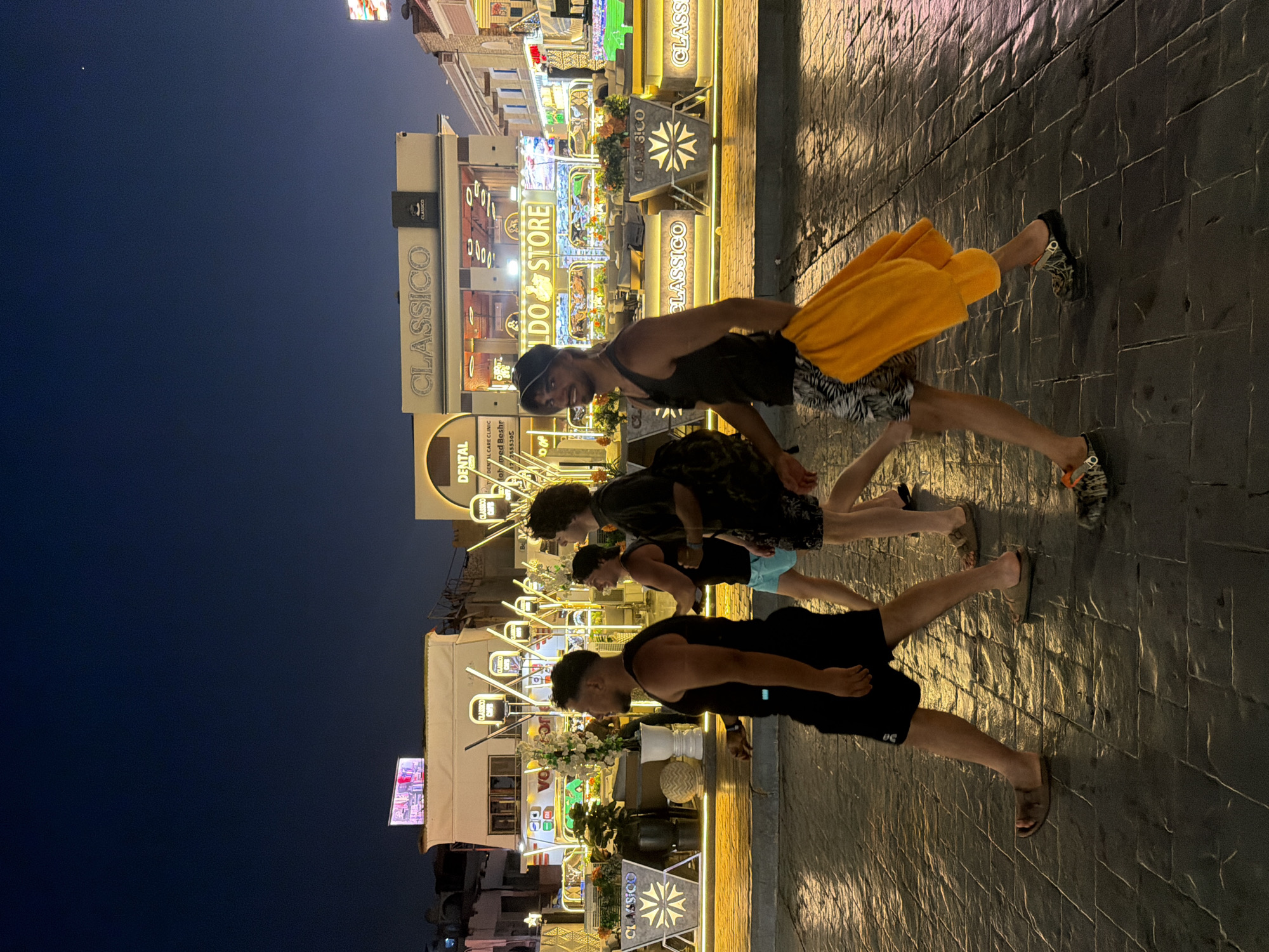 The image depicts a nighttime scene in Sharm El Sheikh, Egypt. Four young men are walking on a wet, cobblestone sidewalk. The man in the foreground, on the right, is wearing a black tank top, patterned shorts, and dark clogs with orange accents. He is carrying a bright orange towel over his shoulder. To his left, another man in a black tank top and blue shorts is walking, partially obscured. The third man, closer to the left edge of the frame, wears a black tank top and black shorts. A fourth man, most to the left, is also wearing a black tank top and black shorts.
The background features a brightly lit pedestrian street with several shops and establishments. Prominent signs include "CLASSICO," "DENTAL CARE CLINIC," and "CHILDO STORE." The architecture appears to be modern with some ornate decorative elements, including illuminated planters with flowers and geometric light fixtures. The overall atmosphere suggests a lively commercial area after dark.