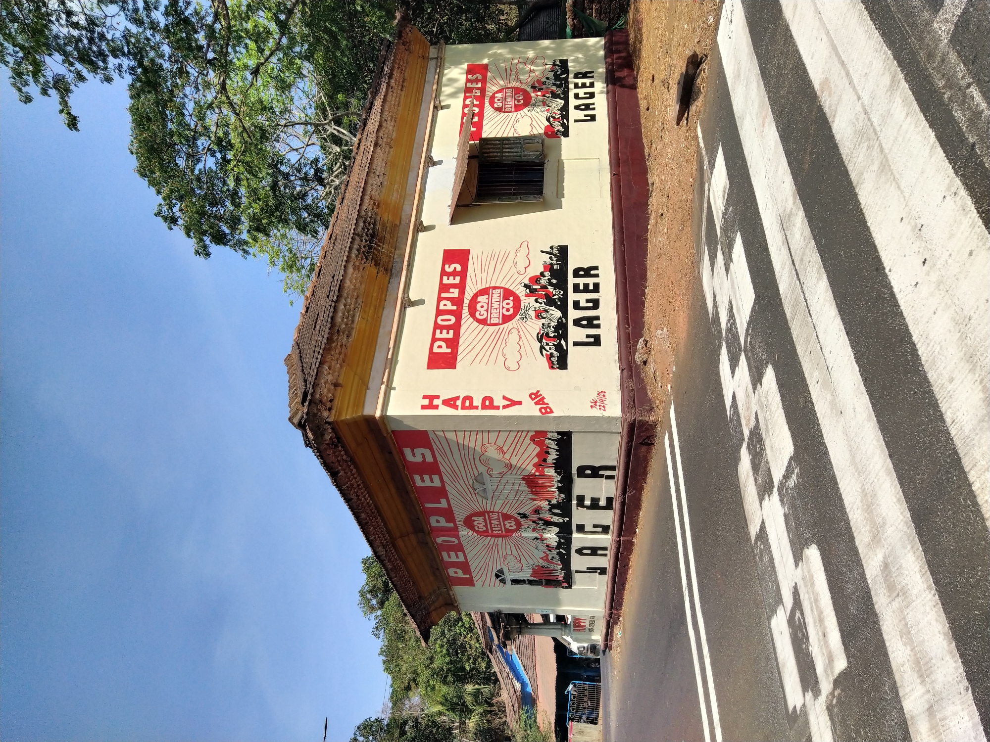 The image shows a low-angle view of a building in Assagao, India, displaying advertising for "PEOPLES GOA BREWING CO. LAGER." The building's exterior is painted in a cream color with red accents for the advertising, which includes an illustration of a sunburst and people. The text "HAPPY BAR" is visible between the two lager advertisements. The building has a tiled roof and a barred window. The image is taken from the street level with part of a zebra crossing visible. Trees are present in the background, with a clear blue sky above.