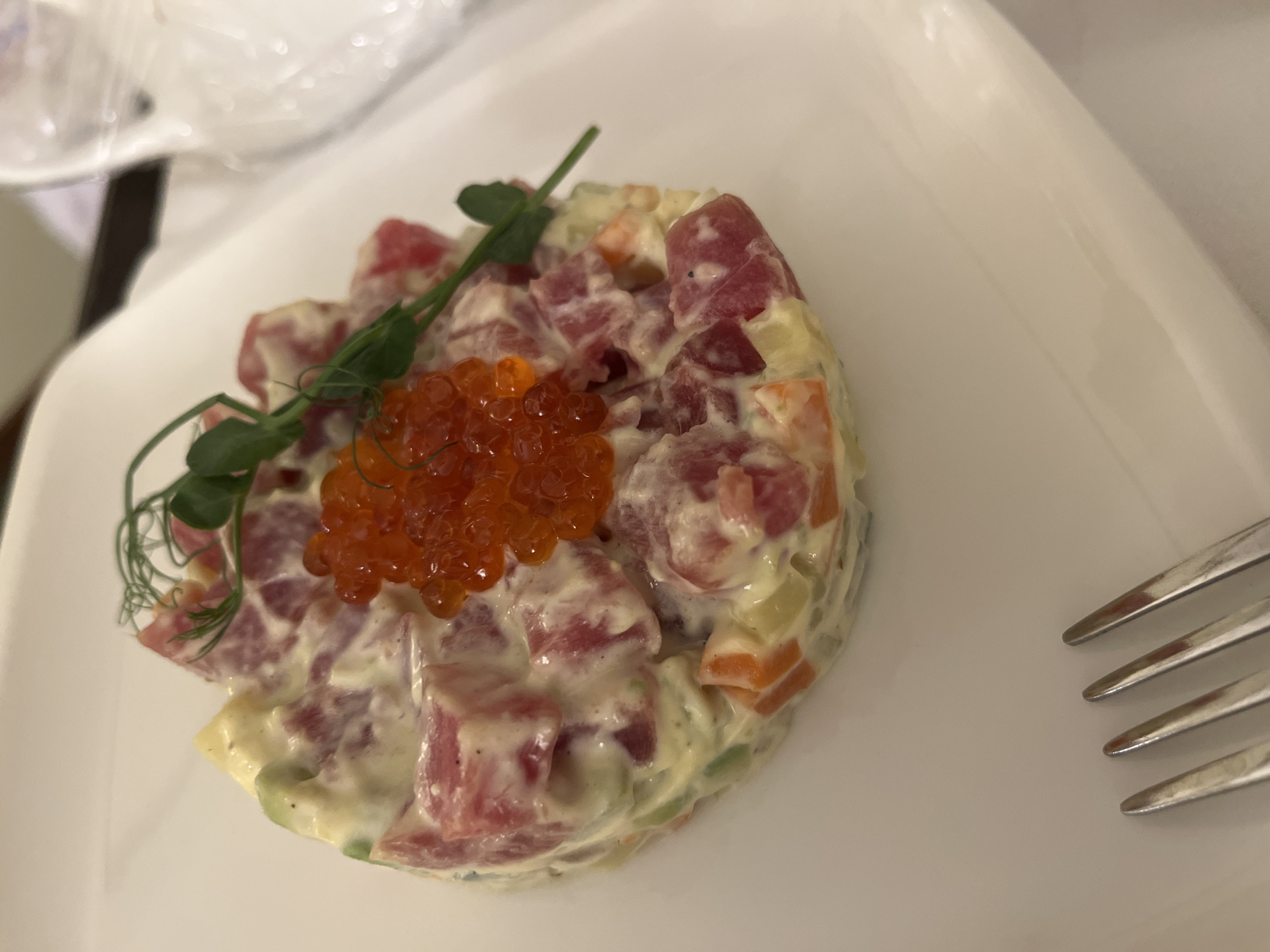 A white plate is holding a salad with cubed ingredients bound by a creamy sauce. The salad is topped with a small mound of orange caviar and a sprig of green sprouts. A fork lies to the right of the plate. The scene appears to be indoors. The location of the image is Moscow, Russia.