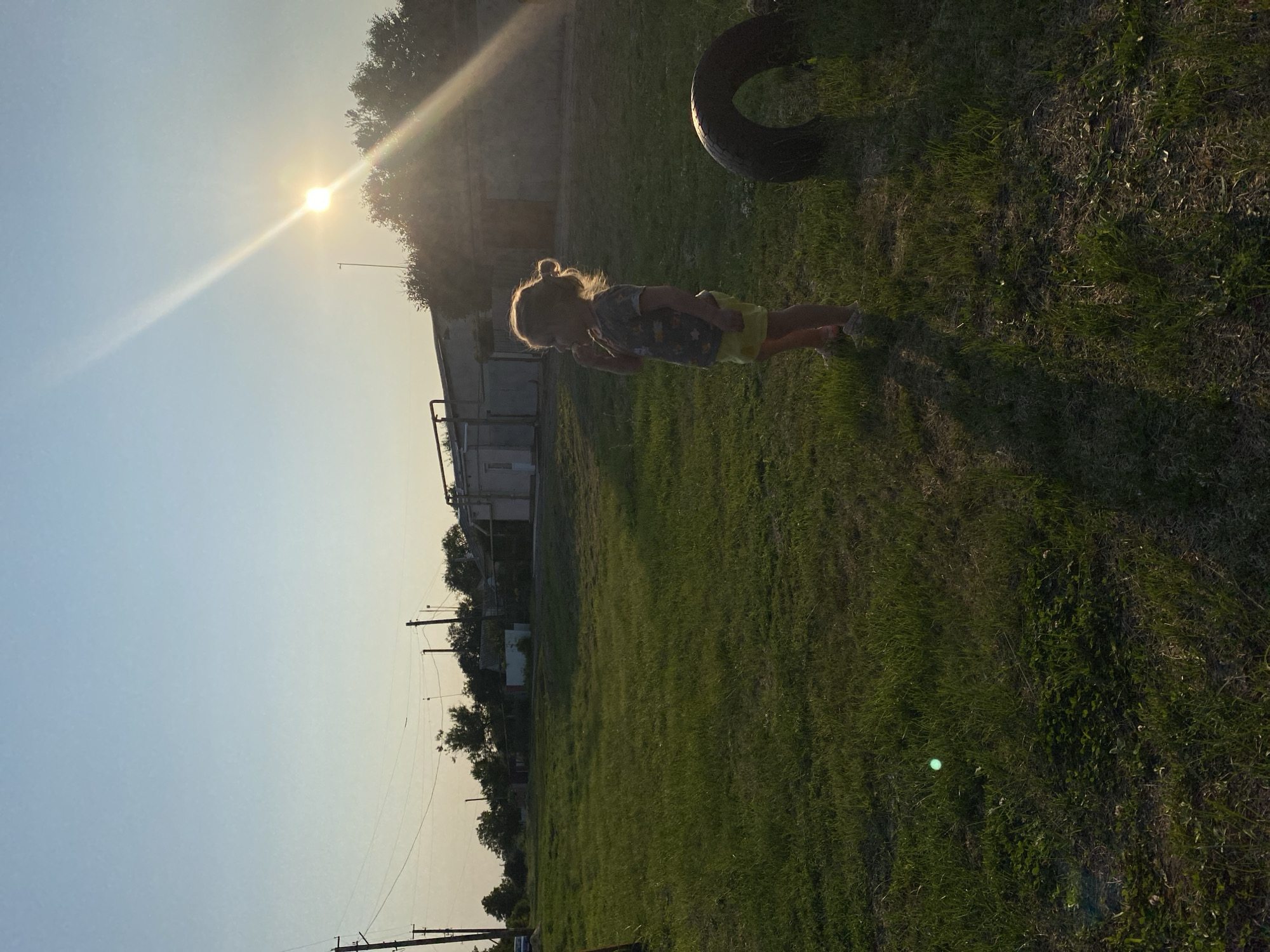 A young girl stands in a grassy field on a sunny day, looking to her left. She has blonde hair pulled up in a bun and is wearing a t-shirt and yellow shorts. The sun is setting, casting a warm glow on the scene and creating a lens flare effect. In the background, there are several buildings, including a light-colored structure with a corrugated roof and some smaller houses. Trees and utility poles are also visible, suggesting a rural or suburban setting. A large tire lies on its side in the foreground to the right of the girl. The overall impression is one of a quiet, relaxed afternoon in the city of Mariupol, Ukraine.